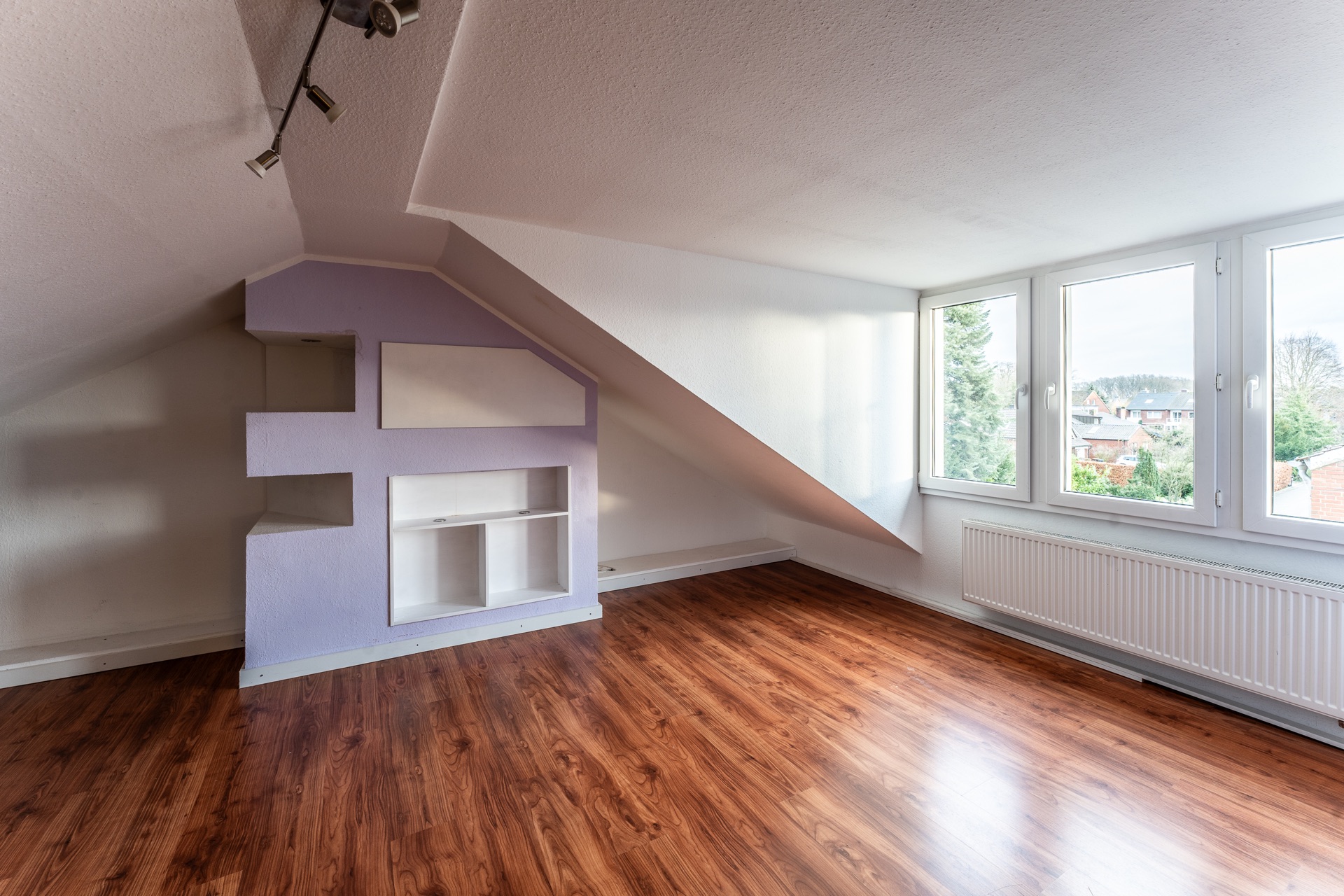 Helles Dachzimmer mit schräger Decke, großem Fenster, Holzboden und eingebautem Regal.