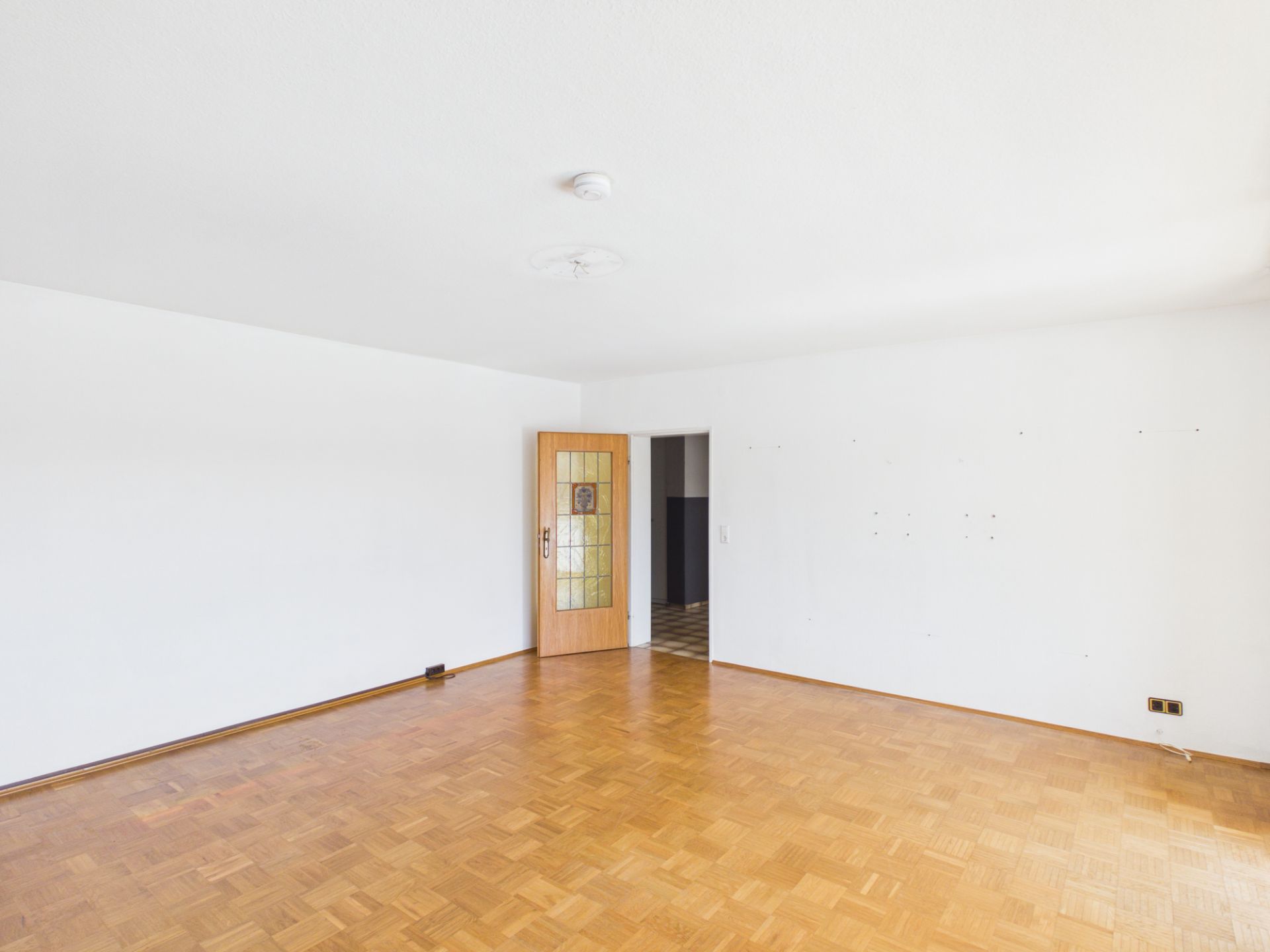 Leerer Raum mit Parkettboden, weißen Wänden und Holztür mit Glaseinsatz.