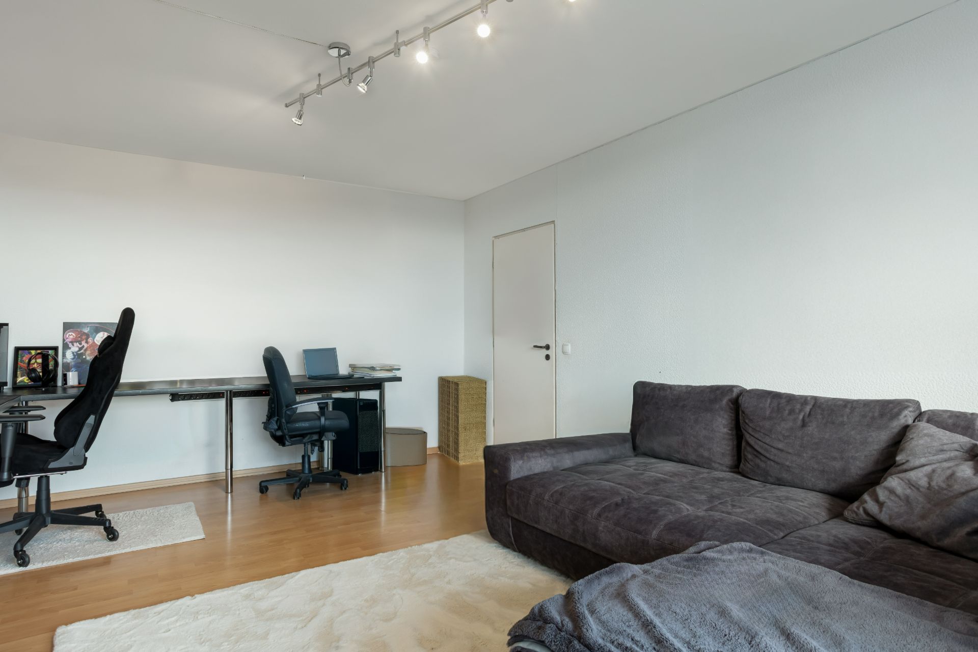 Wohnzimmer mit grauem Sofa, hellem Teppich und zwei Schreibtischen mit Bürostühlen.