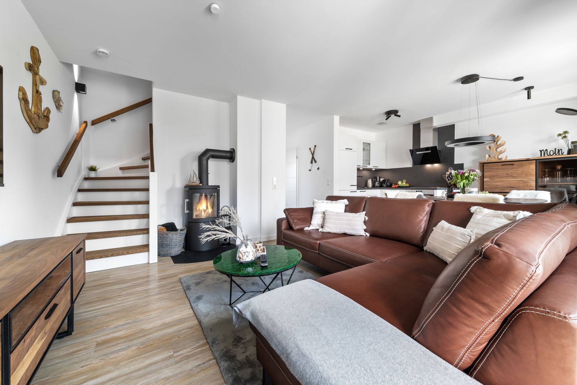 Modernes Wohnzimmer mit braunem Sofa, Kaminofen und offener Küche. Treppe führt nach oben.