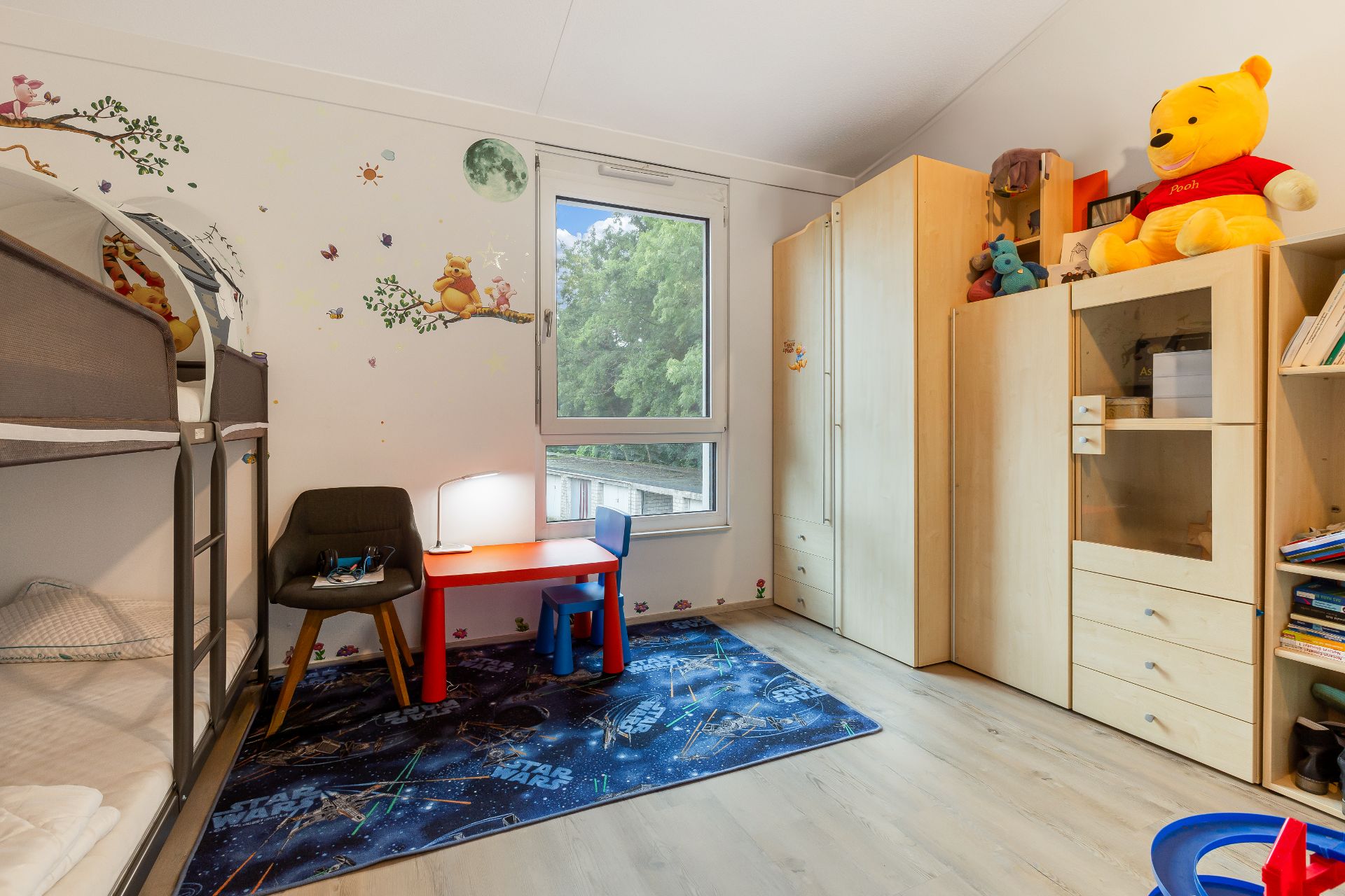 Helles Kinderzimmer mit Etagenbett, Schrankwand, kleinem Tisch und Wanddeko.
