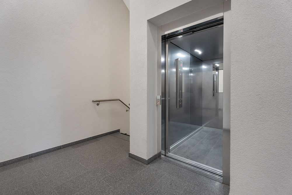 Offener Aufzug neben einer Treppe in einem hellen Flur mit grauem Boden.