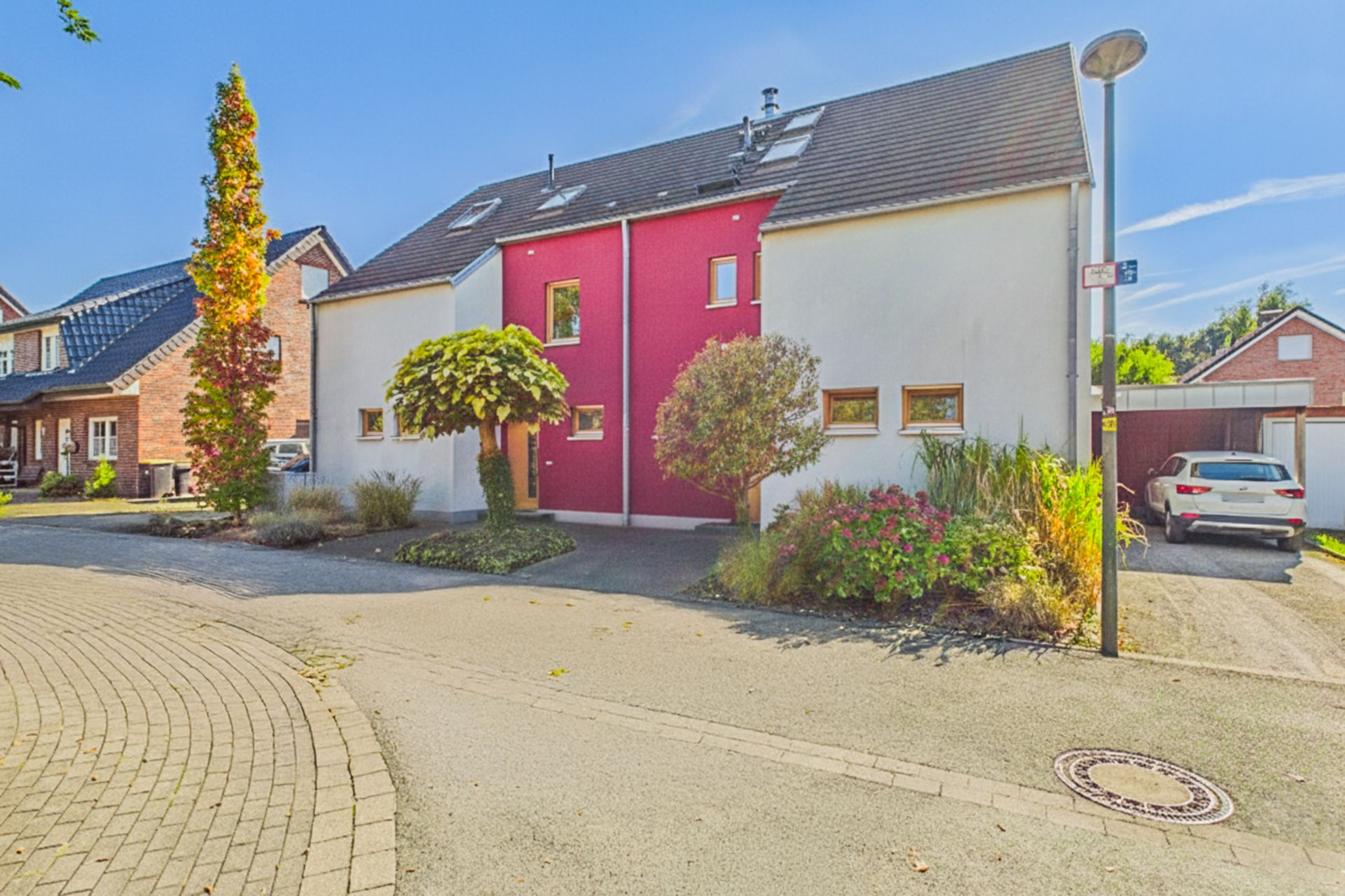 Modernes Wohnhaus mit roter Fassade, kleinen Fenstern und Vorgarten an einer ruhigen Straße.