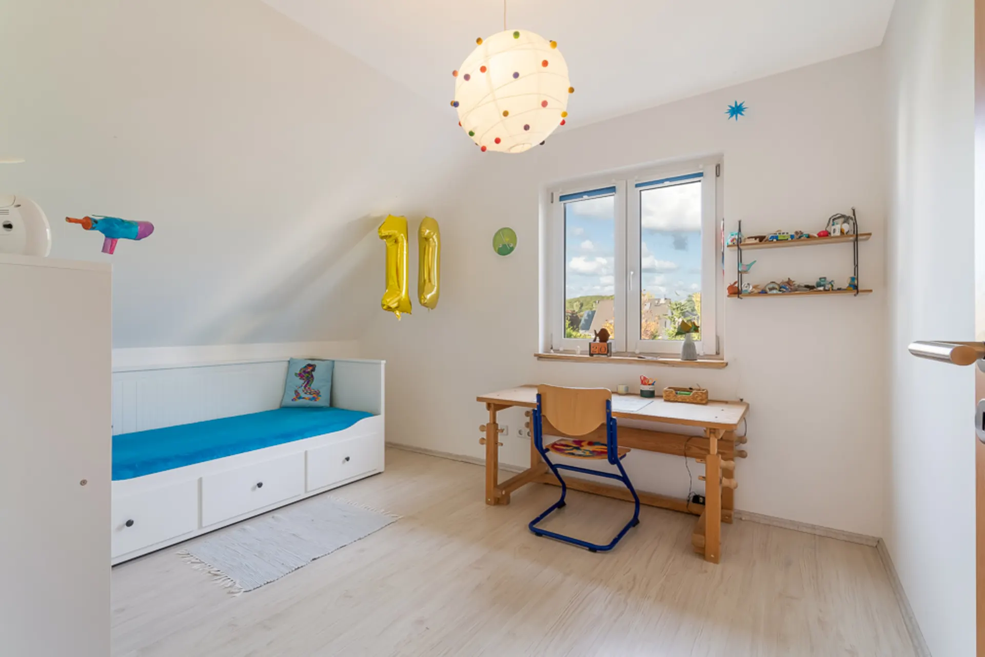 Helles Kinderzimmer mit Bett, Schreibtisch, Regal und einem Fenster mit Ausblick.