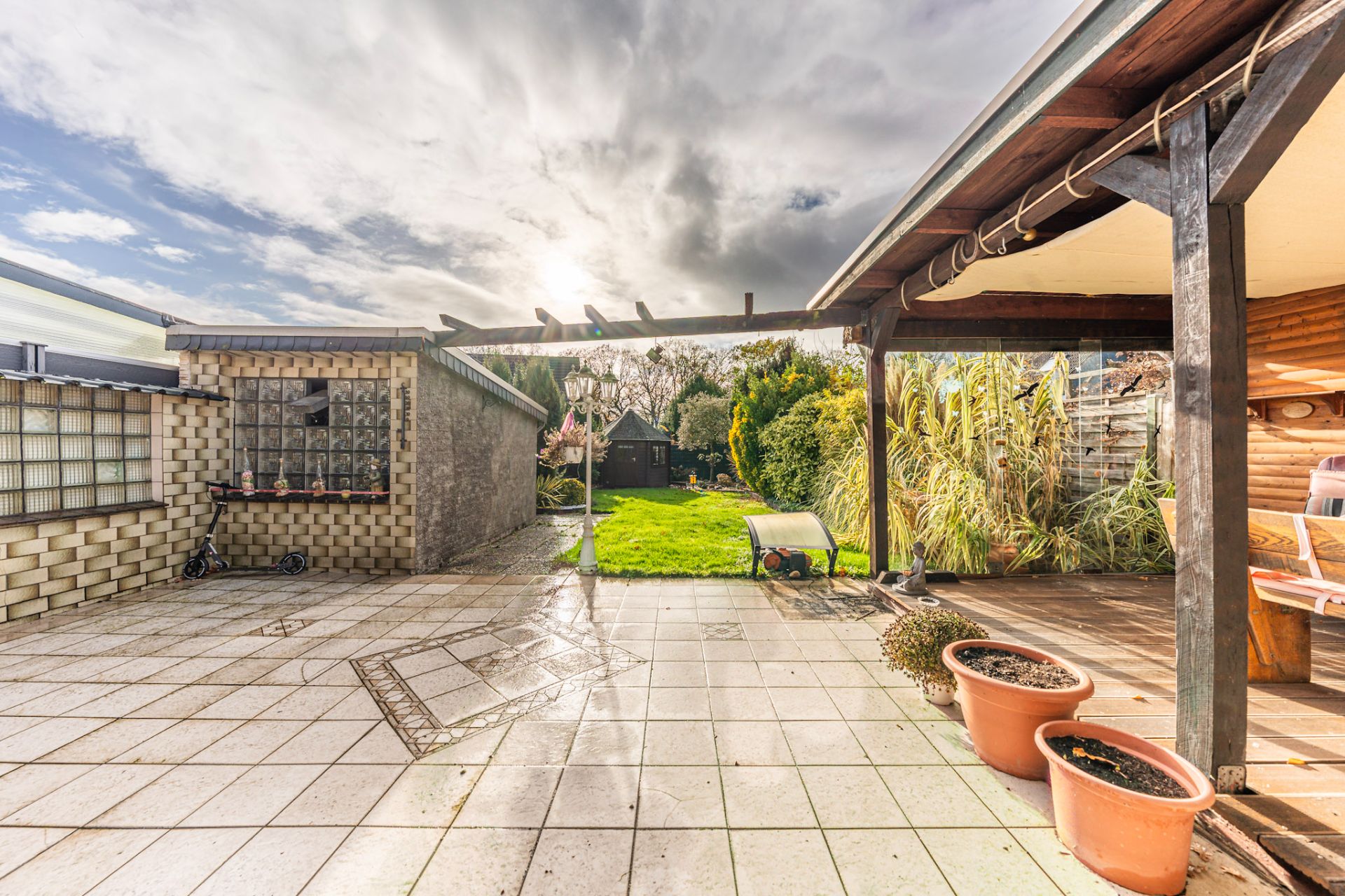 Überdachte Terrasse mit Blumentöpfen, Sitzbereich und Blick auf Garten mit Rasen und Schuppen.