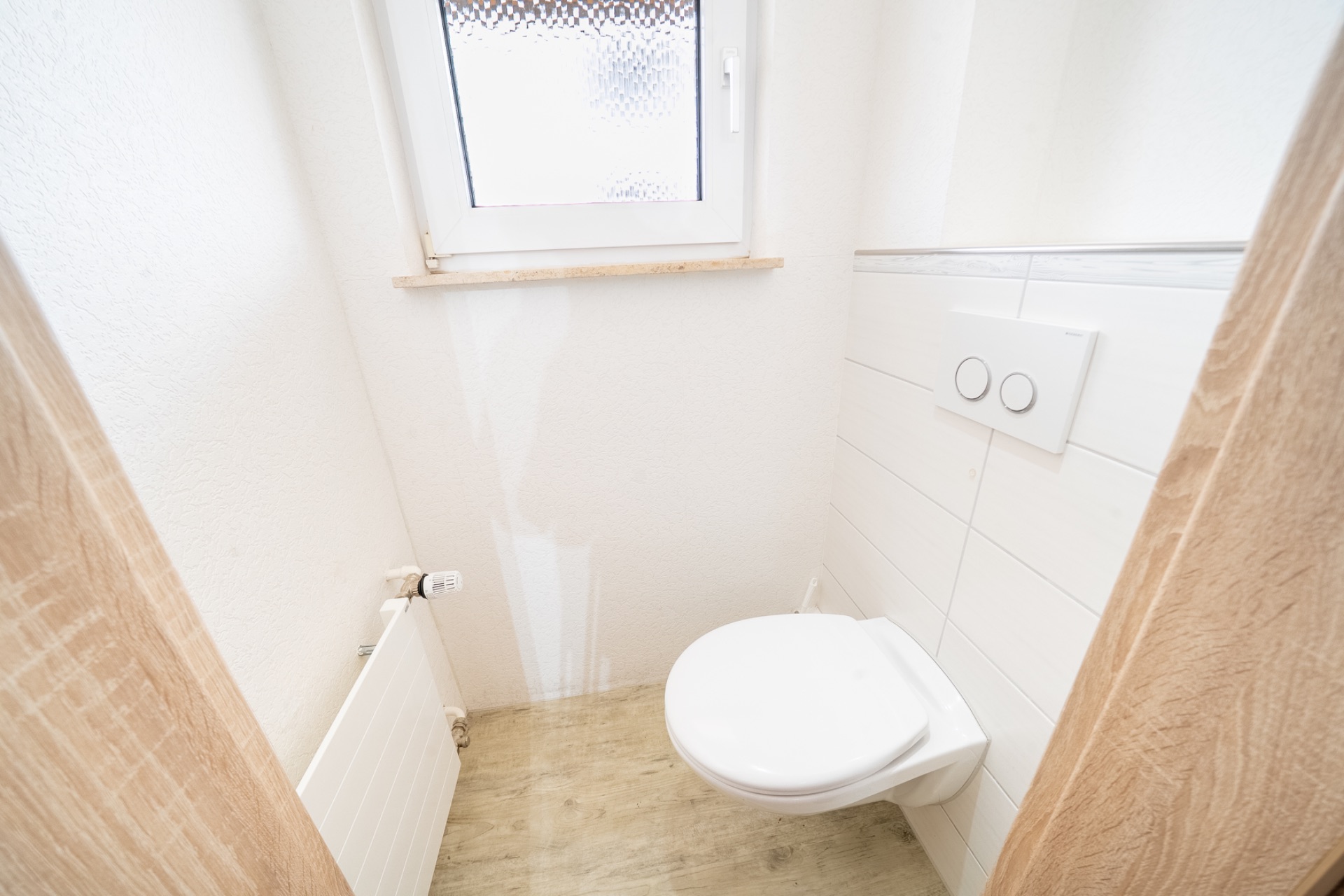 Kleines WC mit wandhängender Toilette, Fenster, Heizung und weißen Fliesen.