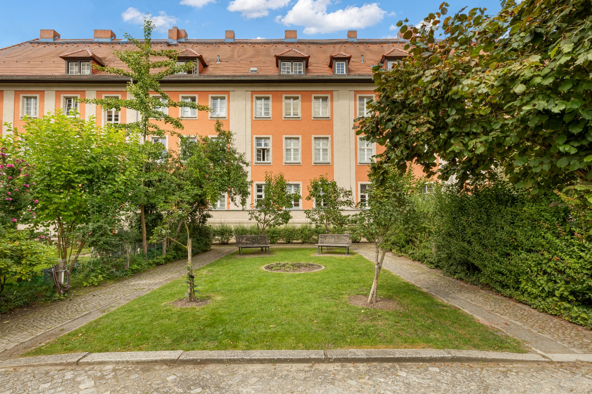 Gepflegter Garten mit Bänken vor einem mehrstöckigen Wohnhaus mit orangener Fassade.