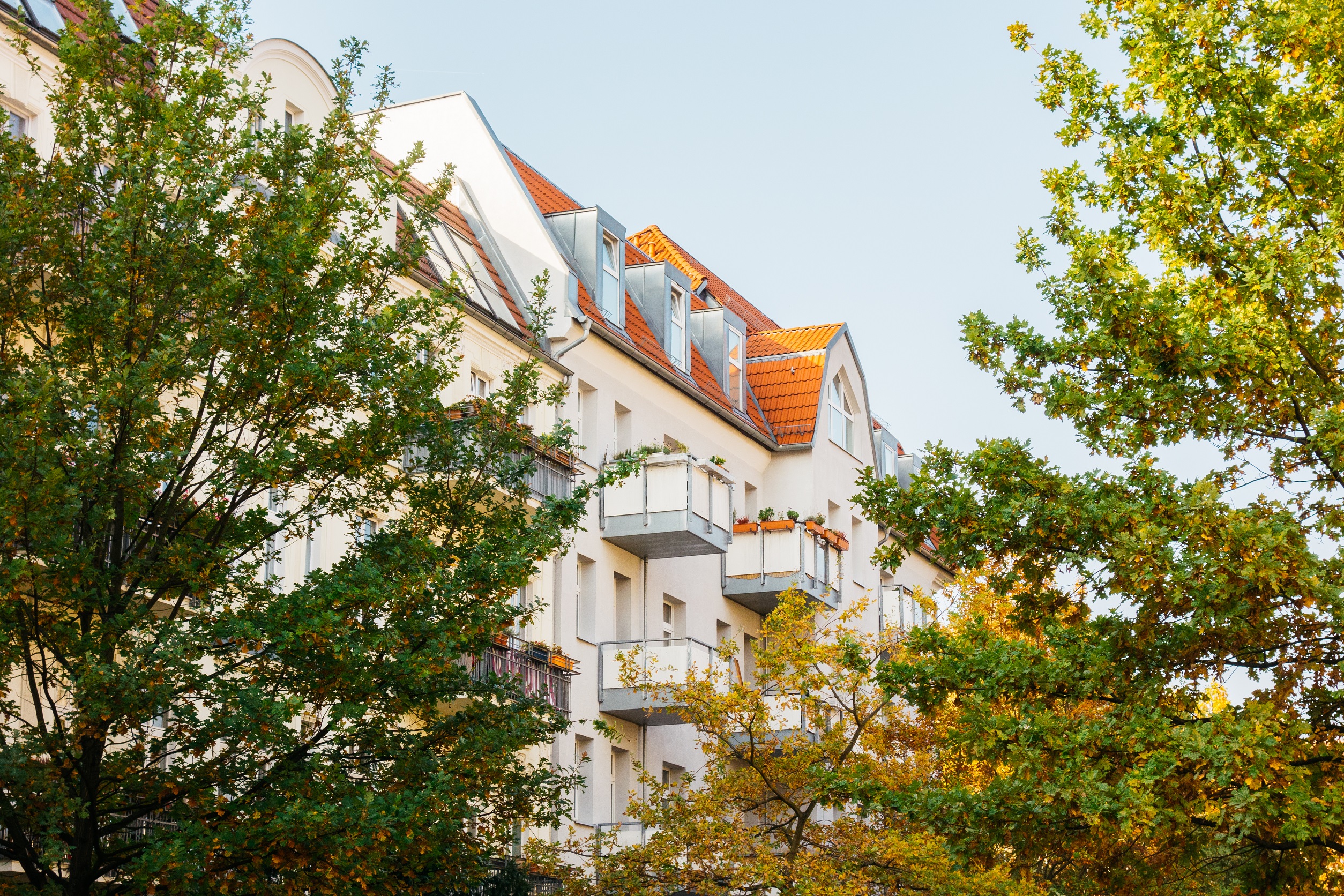 Wohnhaus mit Balkonen und roten Dachziegeln, umgeben von grünen Bäumen.
