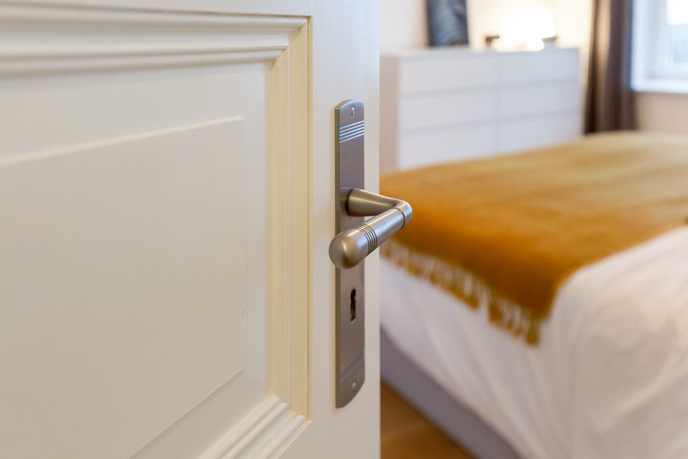 Geöffnete Zimmertür mit Blick auf ein Bett mit gelber Decke und eine weiße Kommode im Hintergrund.