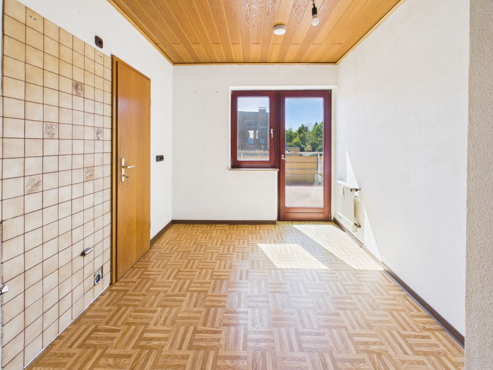 Leerer Raum mit Holzdecke, Parkettboden, Fliesenwand, Tür und Fenster mit Zugang zum Balkon.