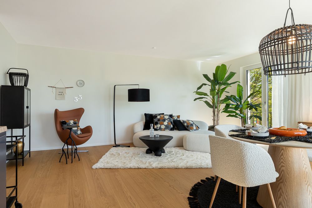 Modernes Wohnzimmer mit weißem Sofa, Sessel, Teppich, Pflanze und Essbereich am Fenster.
