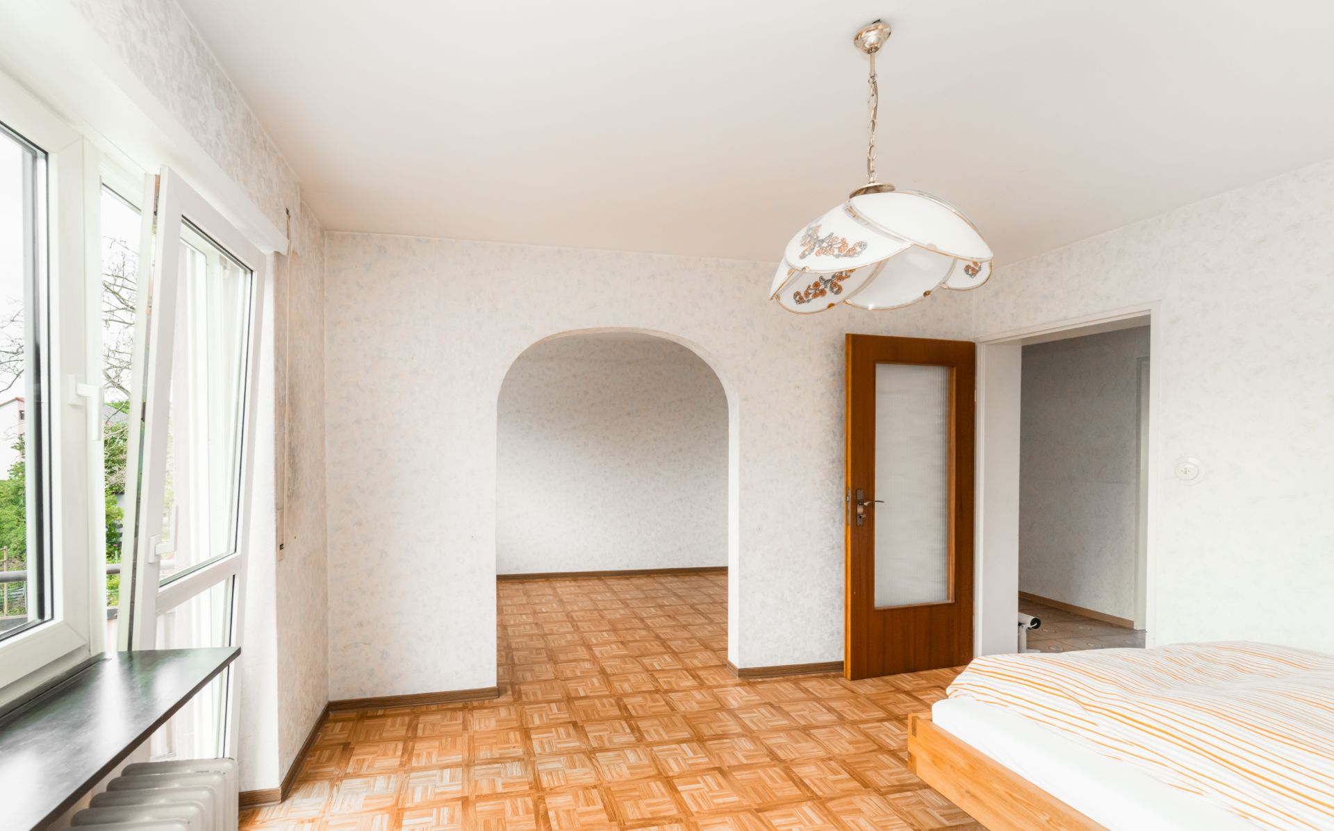 Helles Zimmer mit Bett, großen Fenstern, Holzboden und einem Durchgangsbogen zum nächsten Raum.