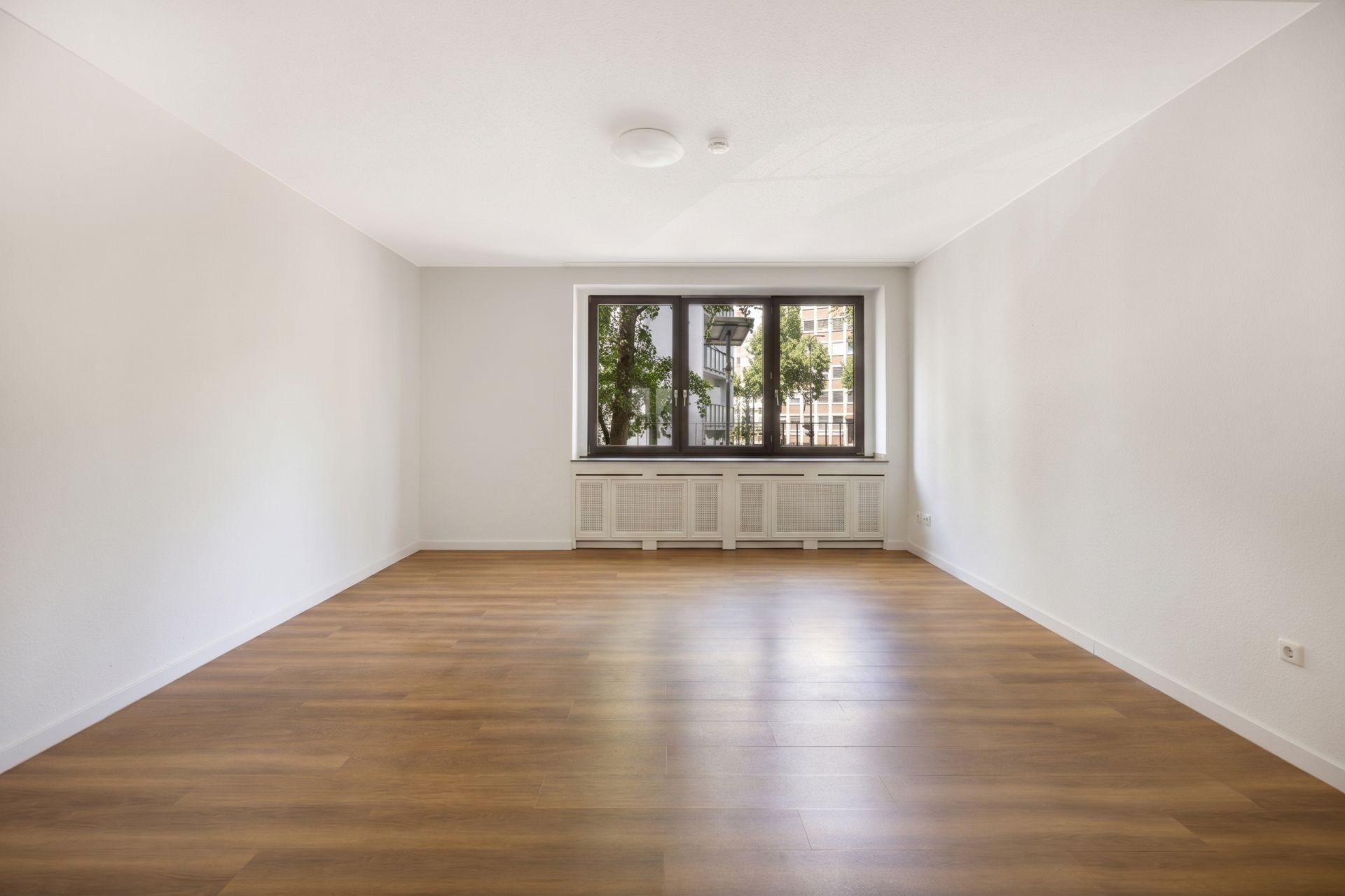 Leerstehendes Zimmer mit Holzfußboden, weiß gestrichenen Wänden und großem Fenster.