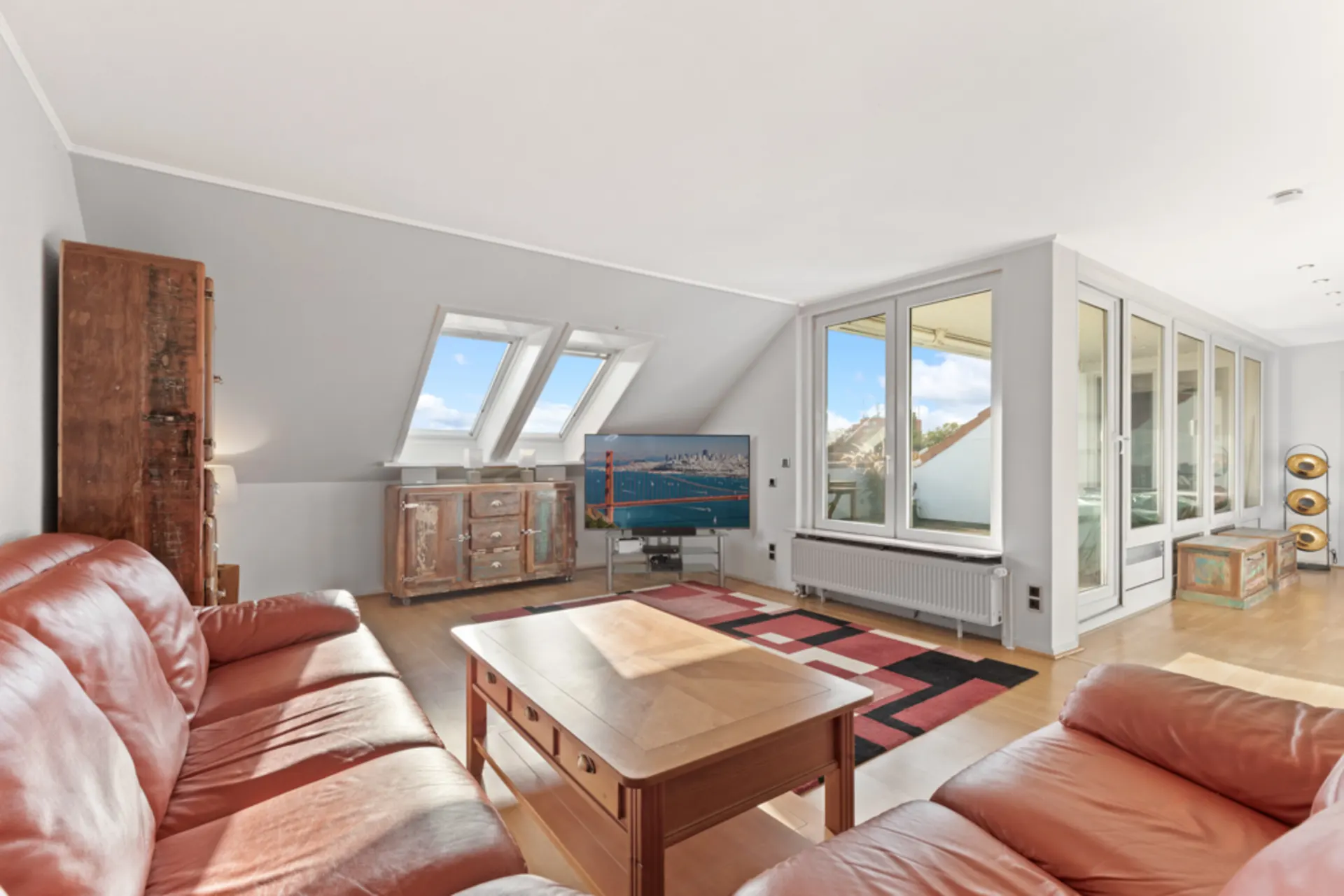Helles Wohnzimmer mit roten Ledersofas, Holzmöbeln, TV und großen Fenstern.
