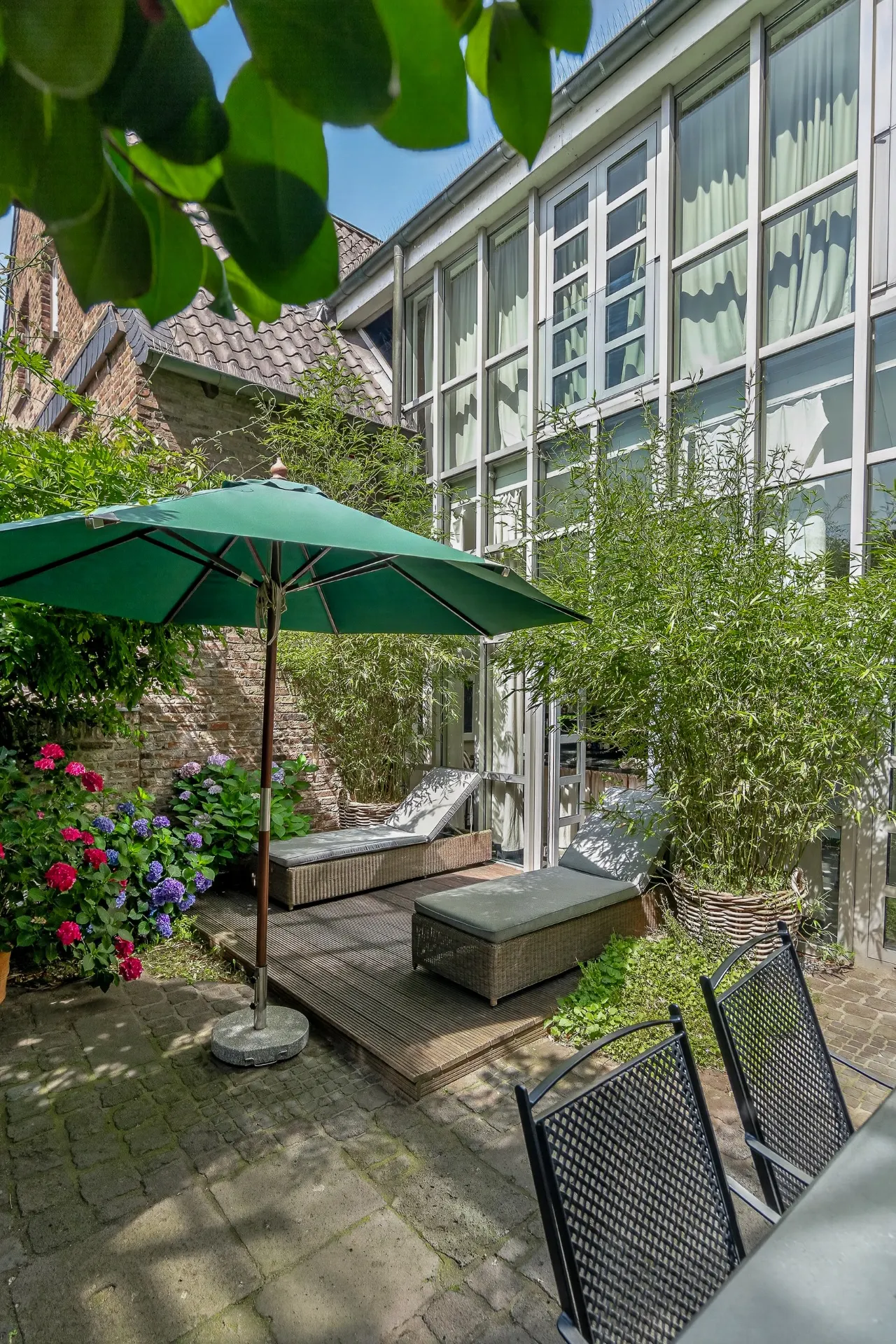 Grüner Innenhof mit Gartenmöbeln, Sonnenschirm, Pflanzen und großen Fenstern eines Wohngebäudes.