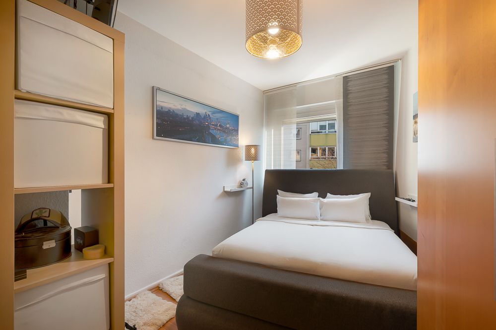 Kleines Schlafzimmer mit Doppelbett, Regal, Lampe und großem Fenster mit Blick nach draußen.