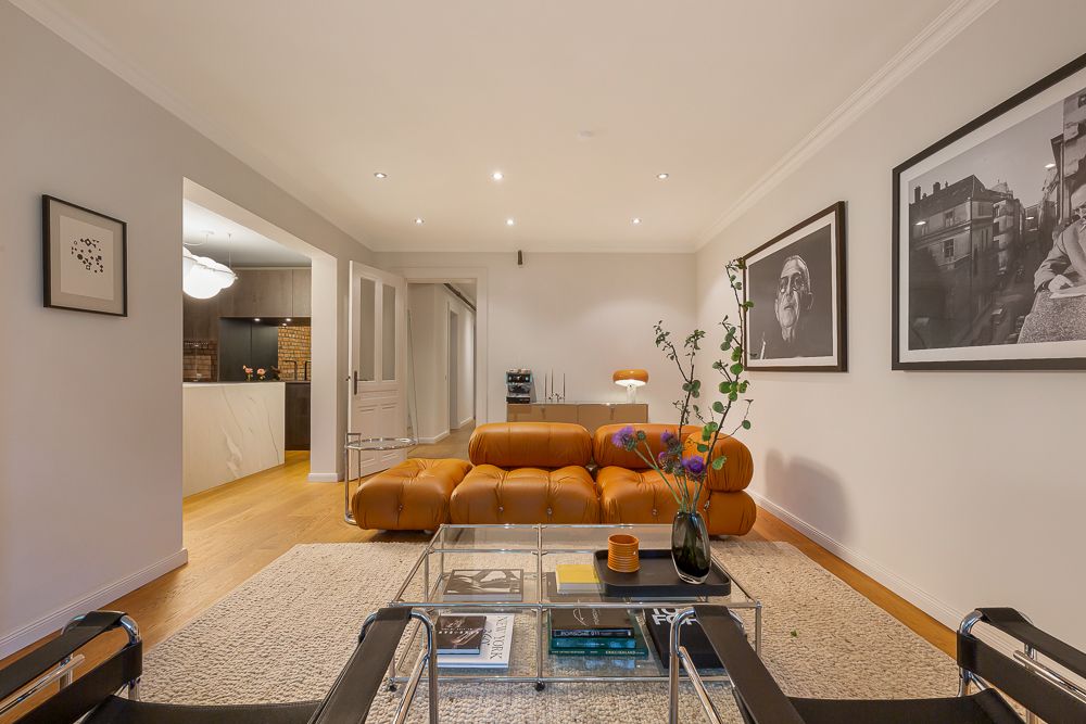 Modernes Wohnzimmer mit braunem Ledersofa, Glastisch und Wandbildern, offener Blick zur Küche.