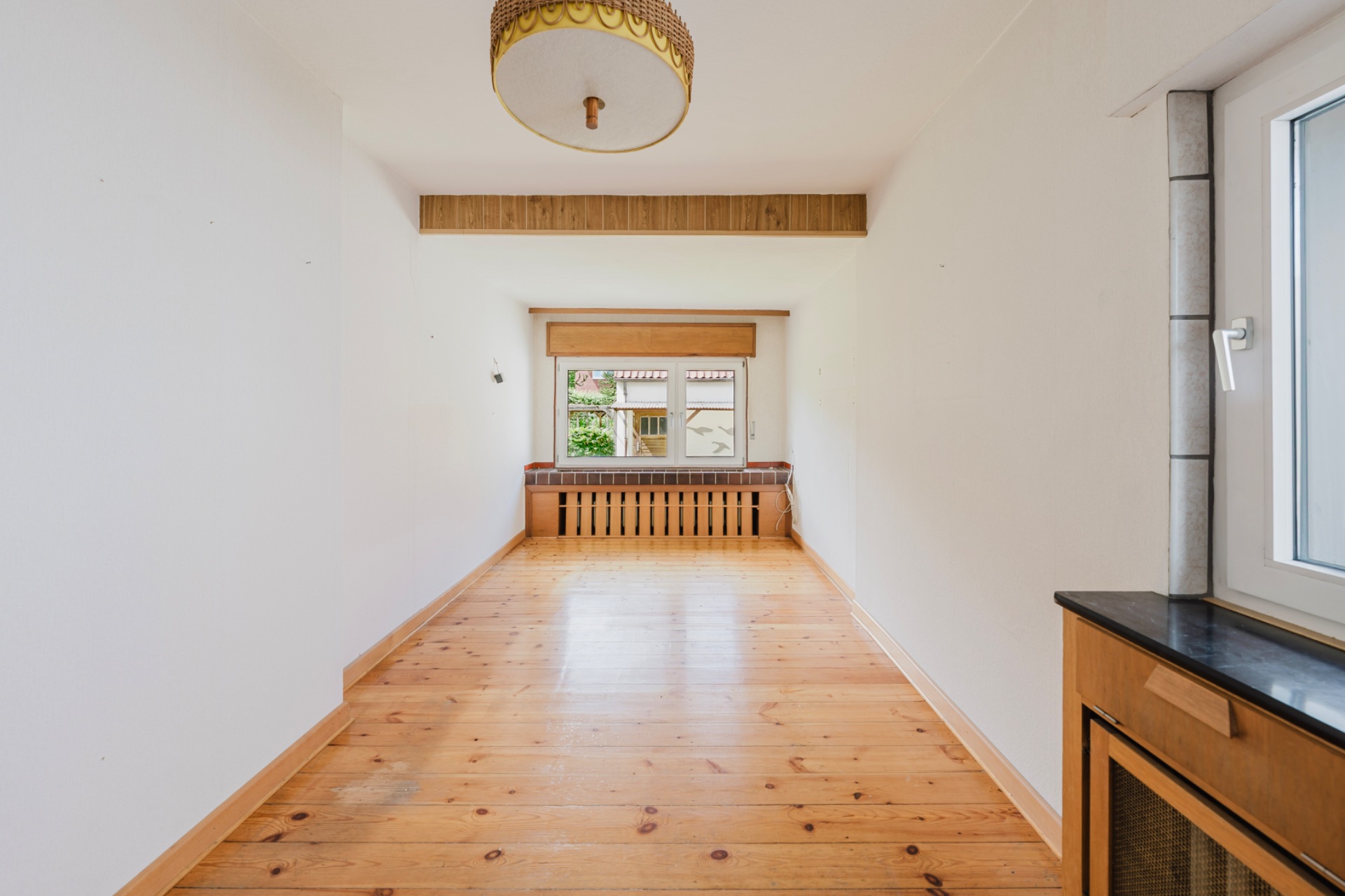Langer, heller Raum mit Holzfußboden, großem Fenster und einer Sitzbank unter dem Fenster.