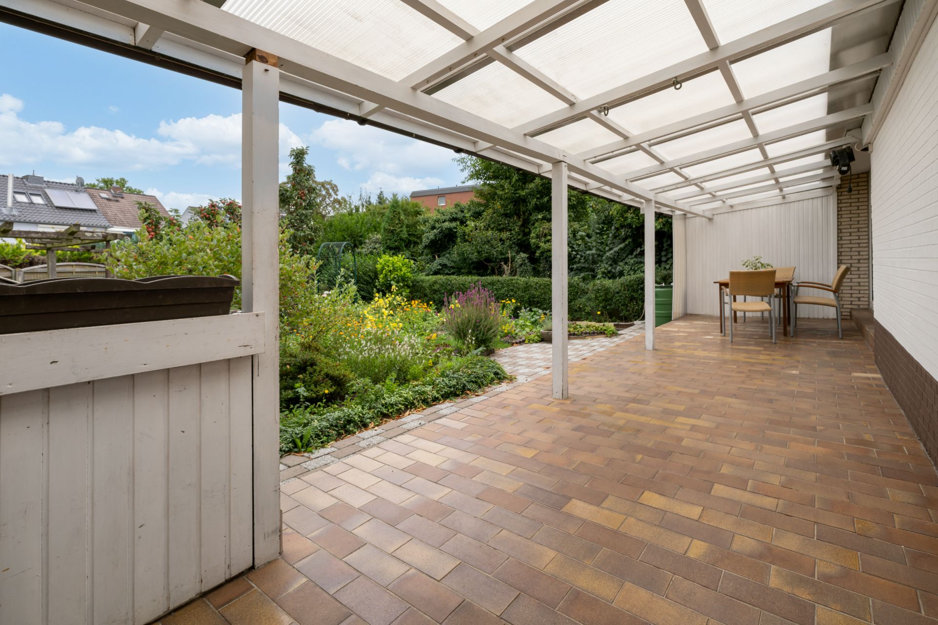 Überdachte Terrasse mit braunen Fliesen und Blick auf einen Garten. Tisch und Stühle im Hintergrund.