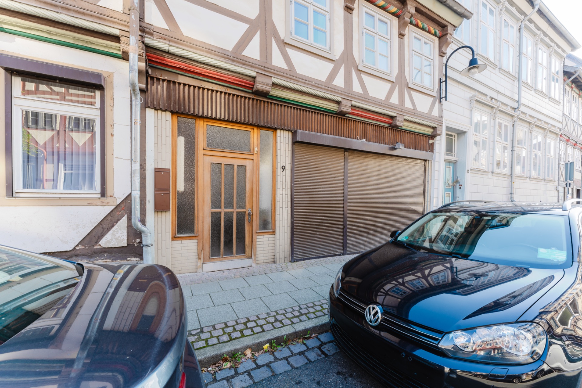 Fachwerkhaus mit Holztür und geschlossenem Rollladen, davor stehen zwei Autos am Straßenrand.