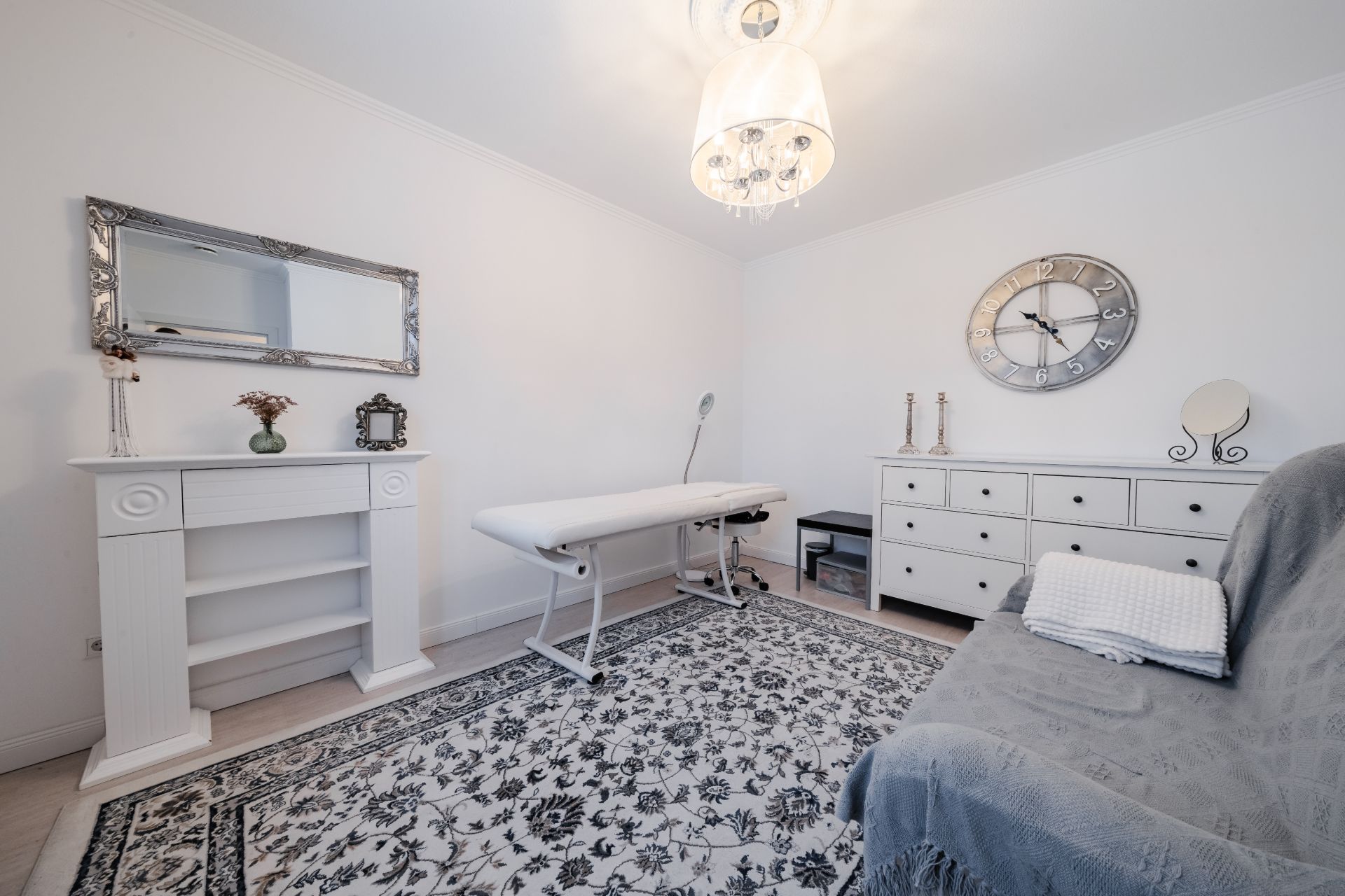 Helles Zimmer mit Massagetisch, Kommode, Spiegel, Sofa und Teppich auf Holzboden.