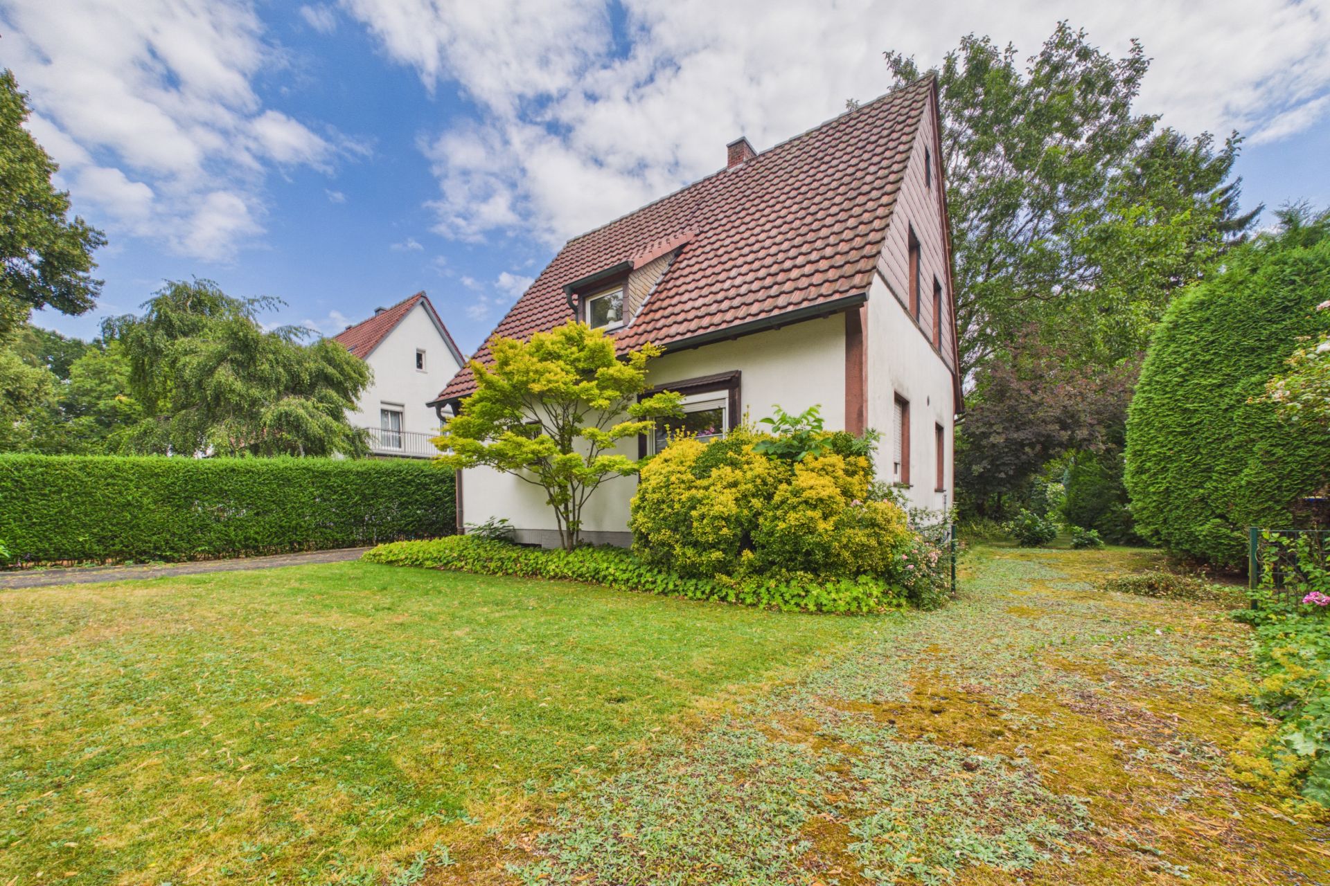 Haus mit rotem Satteldach, Garten und Büschen, umgeben von grüner Hecke.
