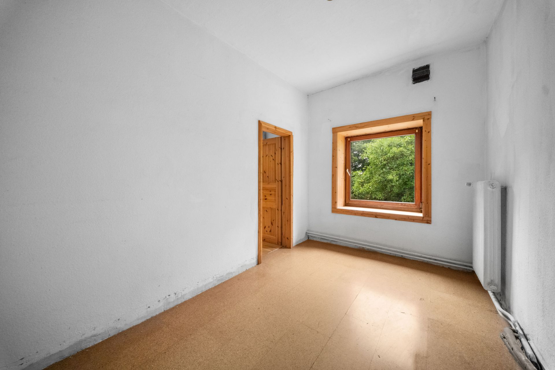 Leeres Zimmer mit großem Fenster, weißer Wand und Holztür, Blick ins Grüne.