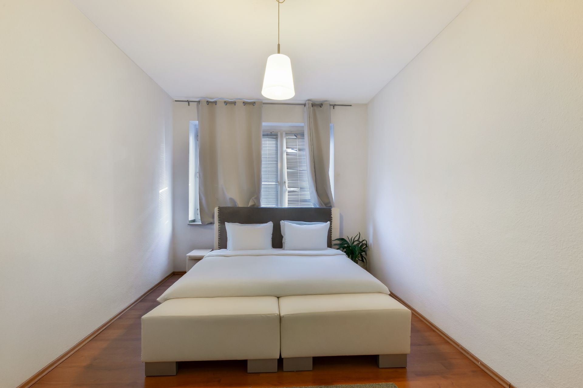 Helles Schlafzimmer mit Doppelbett, zwei Fenstern, Vorhängen und einer kleinen Pflanze am Fenster.