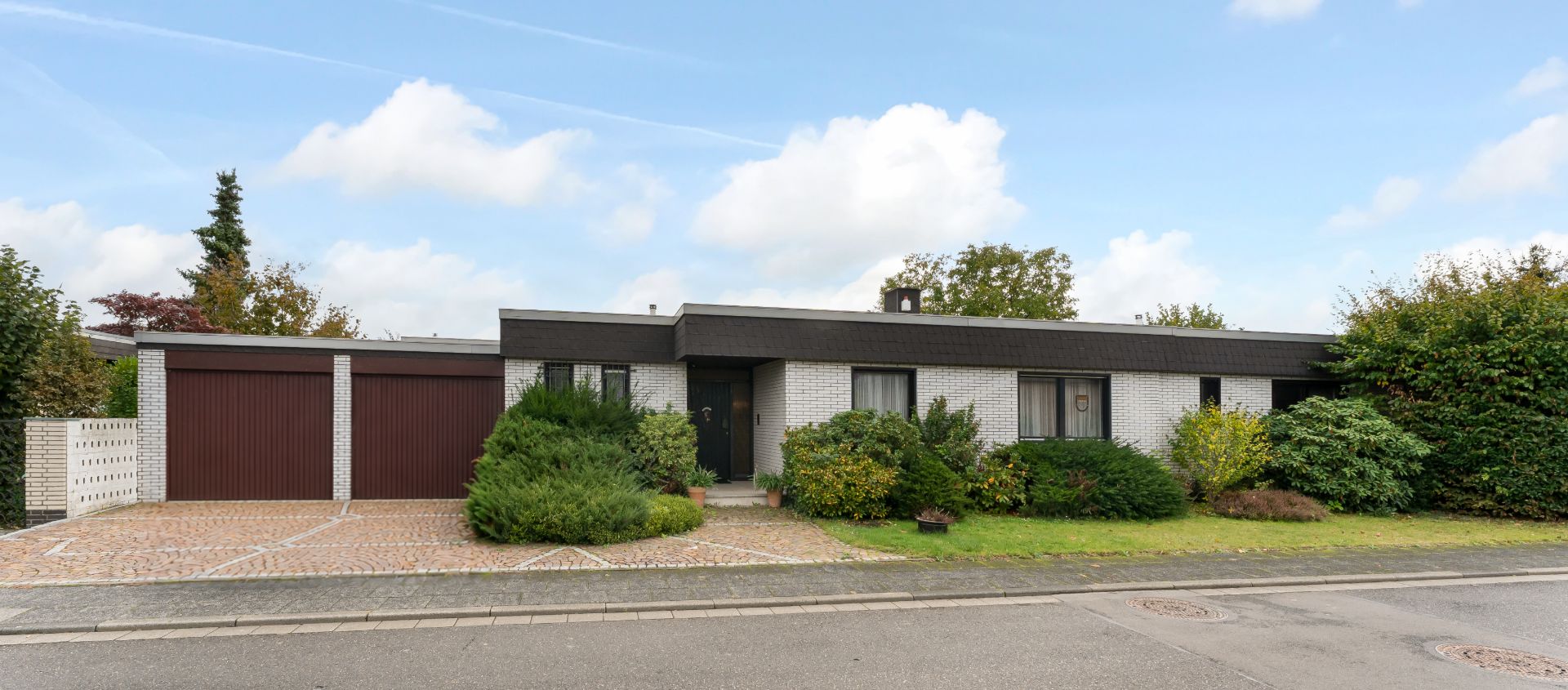Flaches Einfamilienhaus mit Garage, Vorgarten mit Sträuchern und gepflasterter Einfahrt.