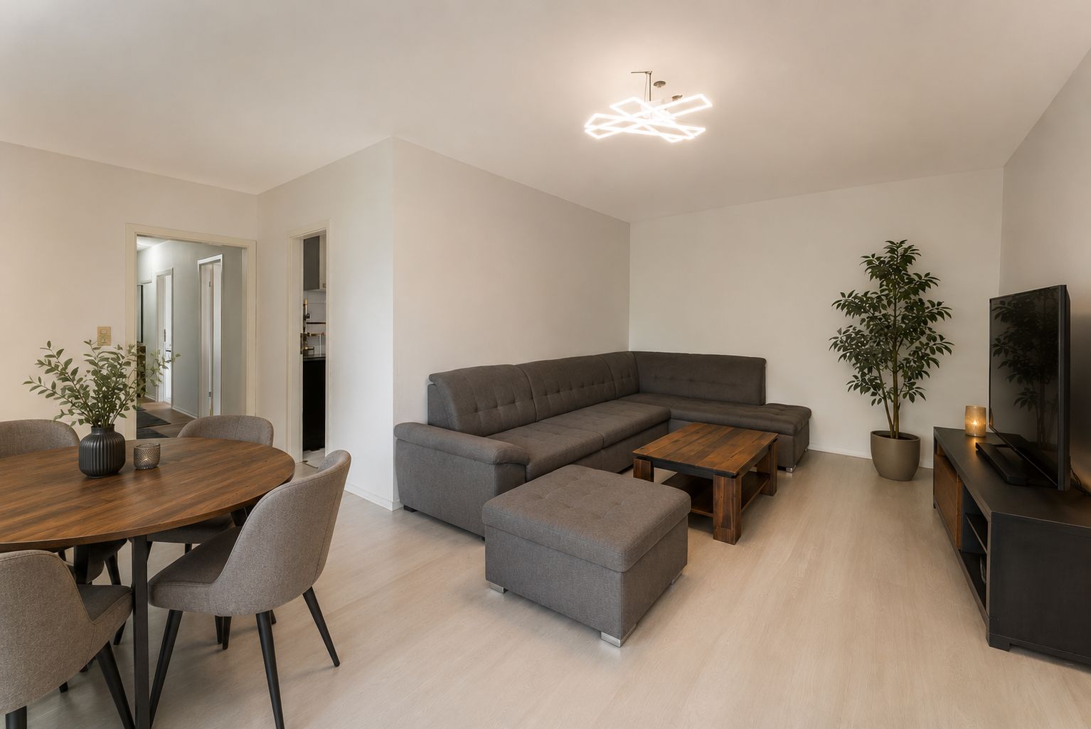 Modernes Wohnzimmer mit grauem Ecksofa, Couchtisch, Esstisch und großem Fernseher.