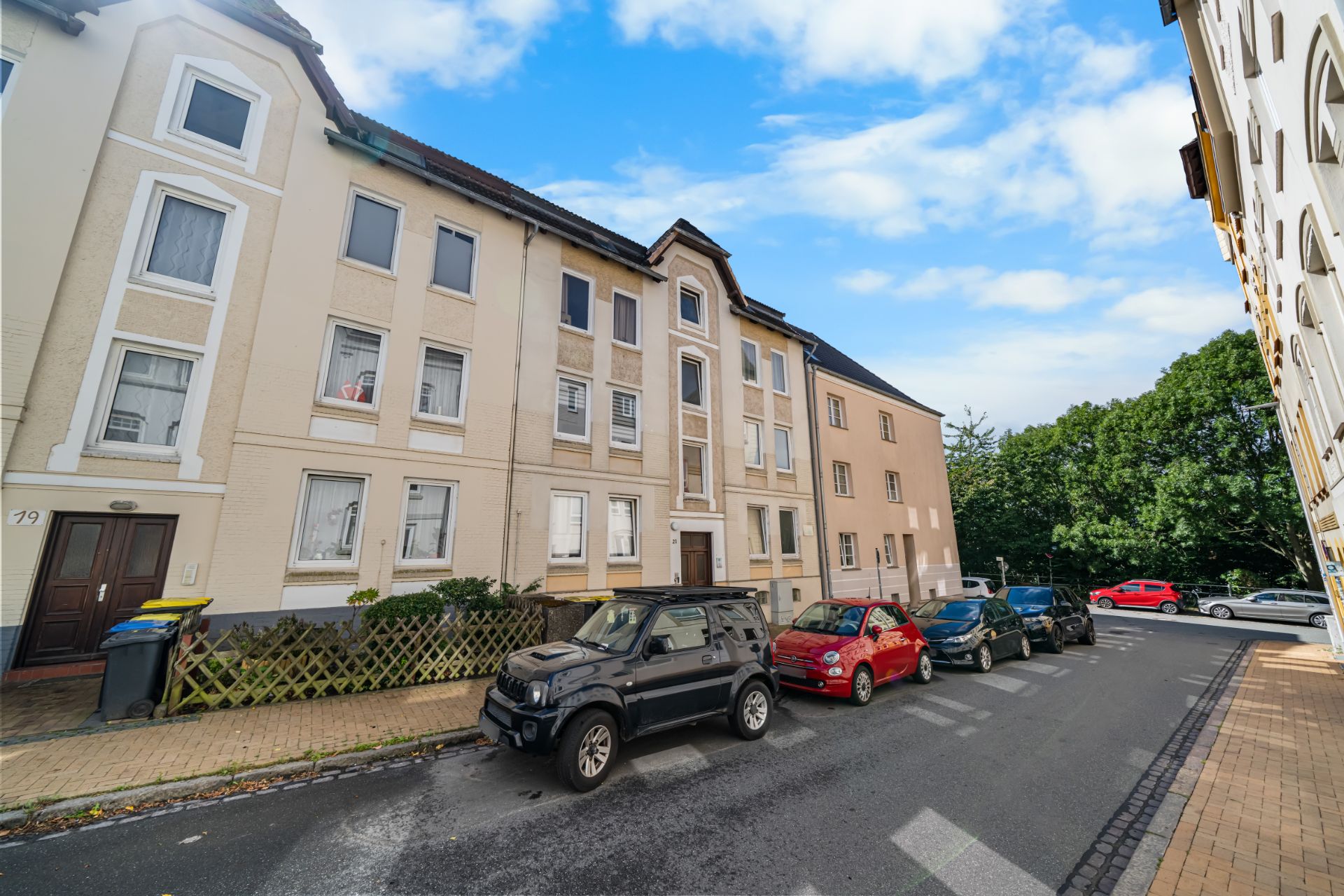 Mehrfamilienhaus mit geparkten Autos an der Straße und blauem Himmel im Hintergrund.