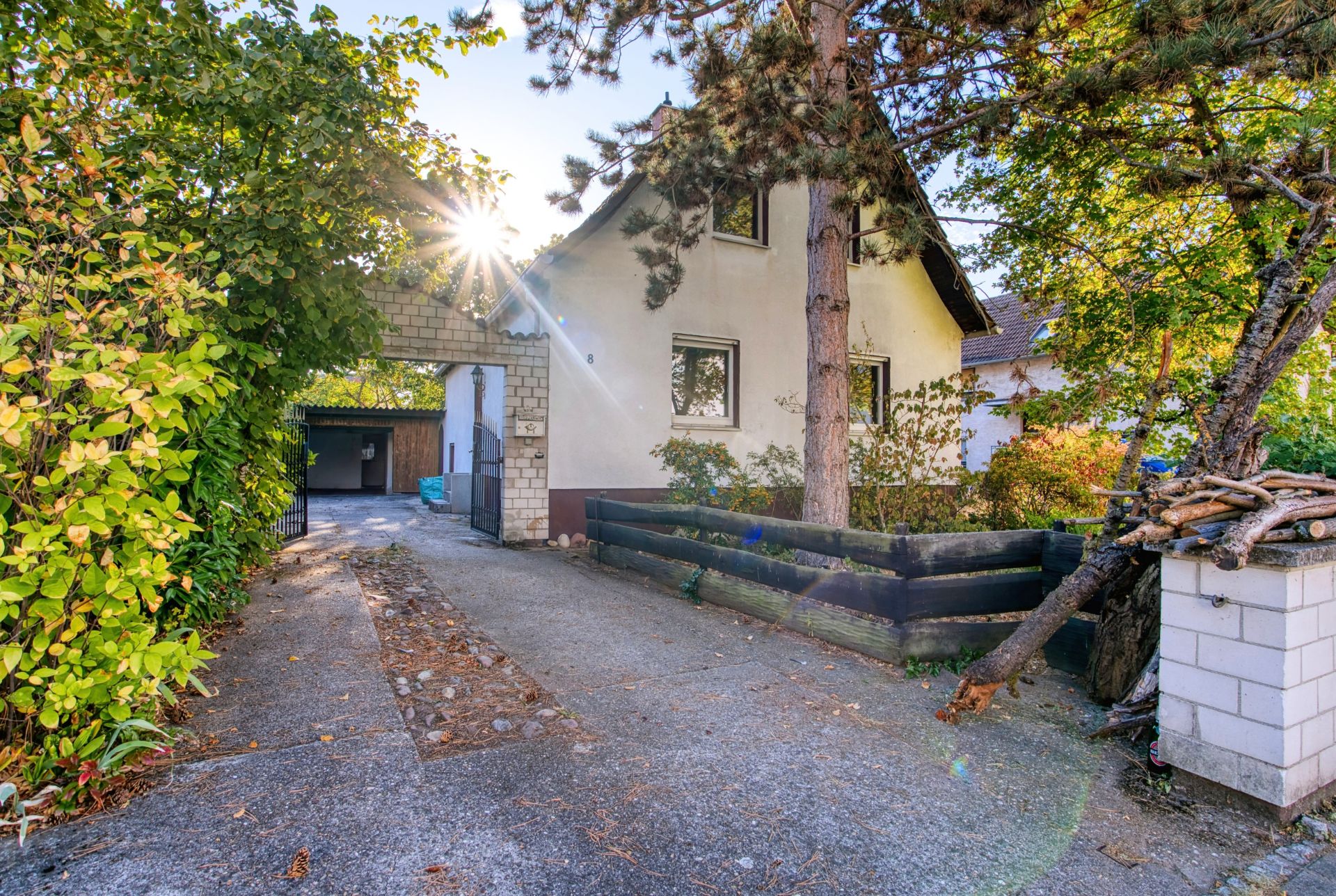 Einfahrt zu einem Einfamilienhaus mit Garten und Bäumen, Sonnenstrahlen im Hintergrund.