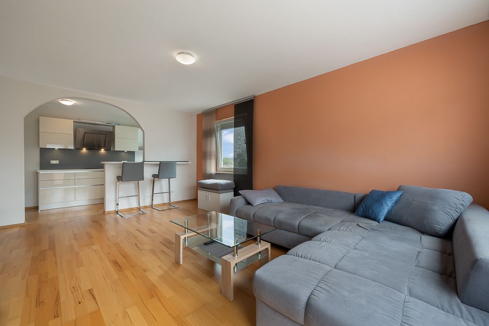 Wohnzimmer mit grauem Ecksofa, Glastisch und offener Küche mit Tresen und zwei Hockern.