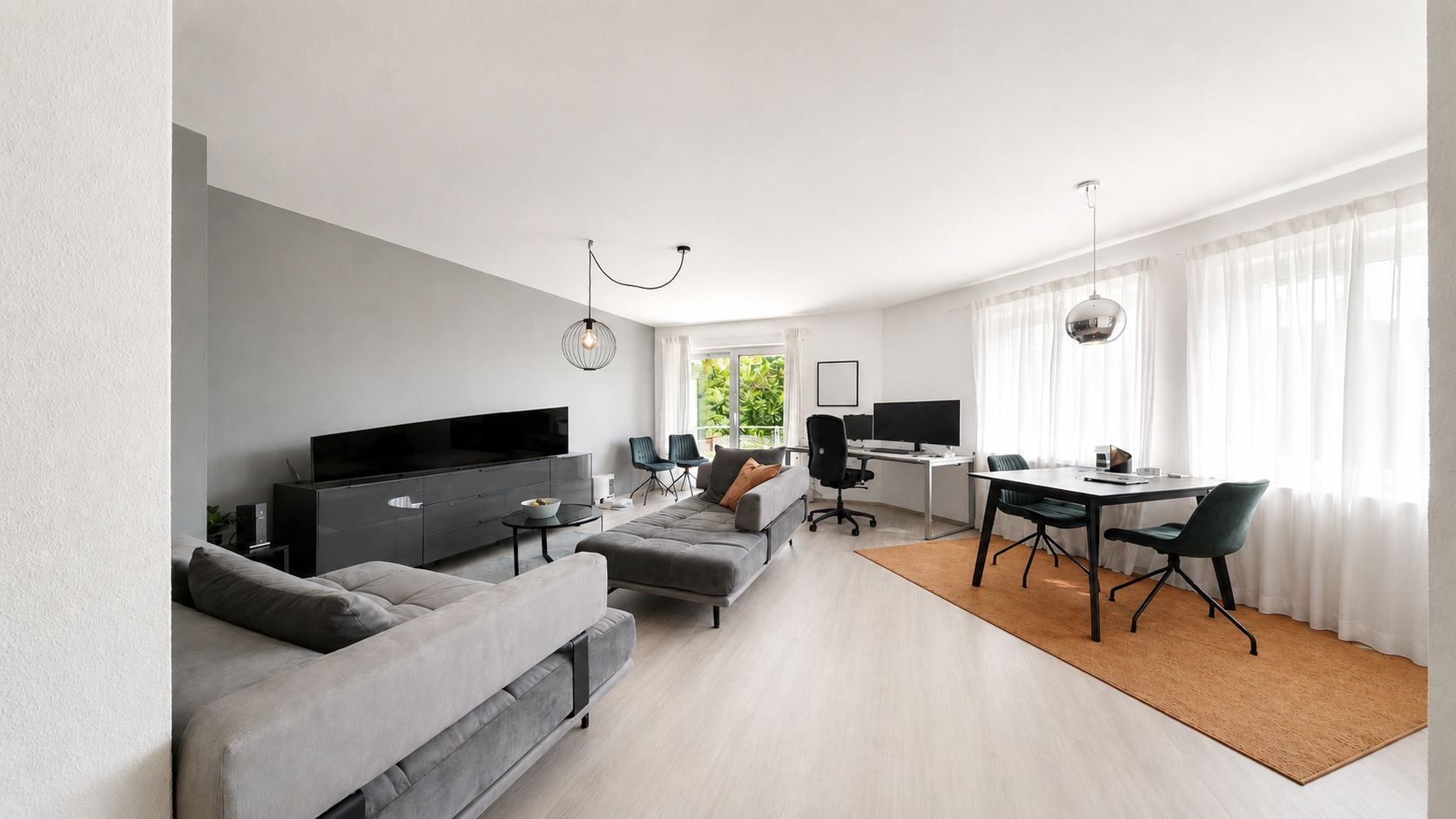 Modernes Wohnzimmer mit grauen Sofas, TV, Schreibtisch und Esstisch an großen Fenstern mit weißen Vorhängen.