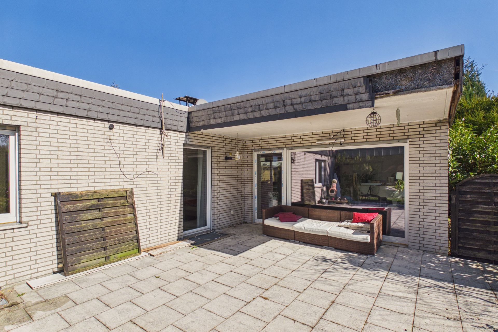 Terrasse mit hellen Fliesen, Sofa, großen Fenstern und weißer Klinkerfassade bei Sonnenschein.