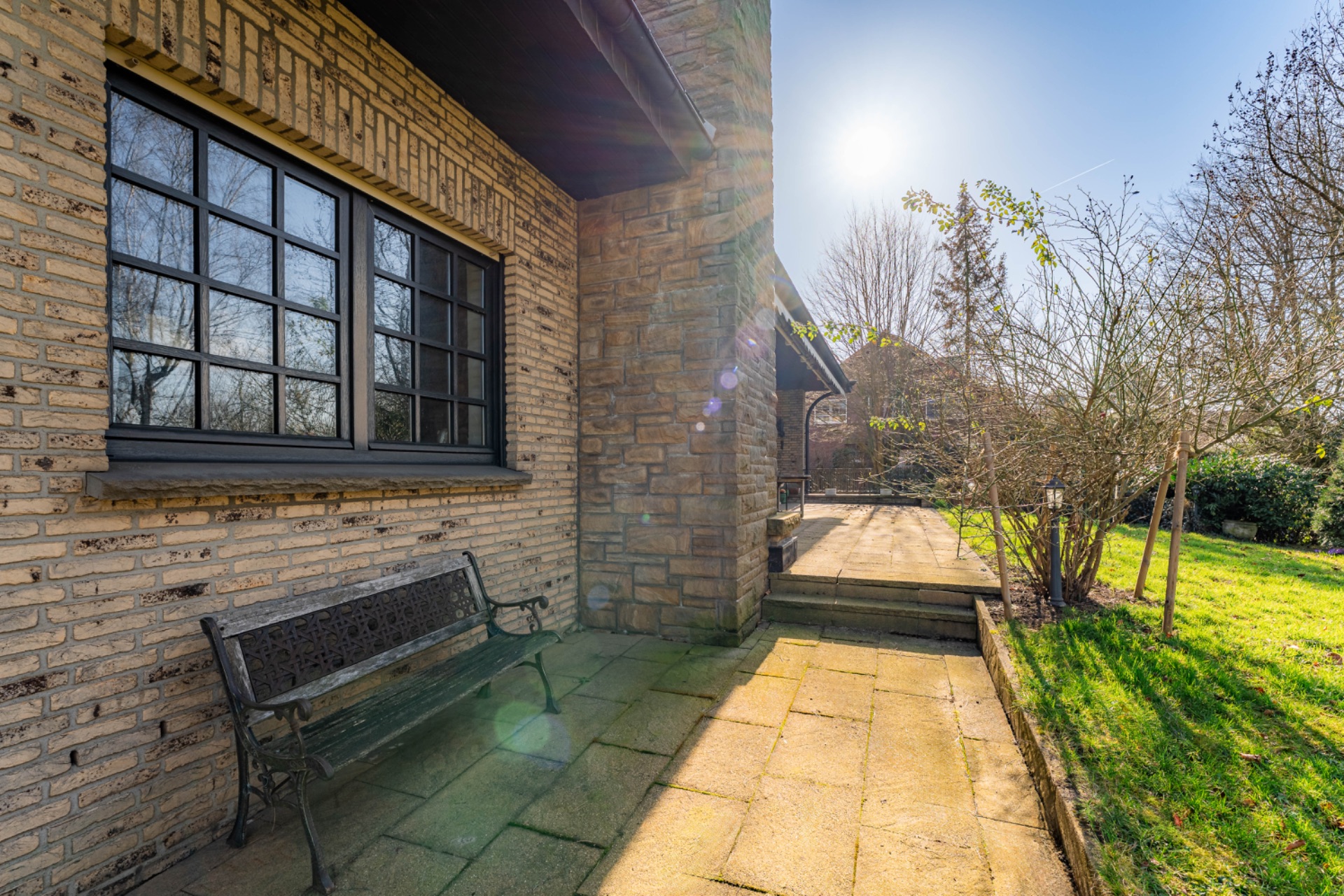 Sonnige Terrasse mit Steinbank, Backsteinfassade und Blick auf Garten mit Bäumen.
