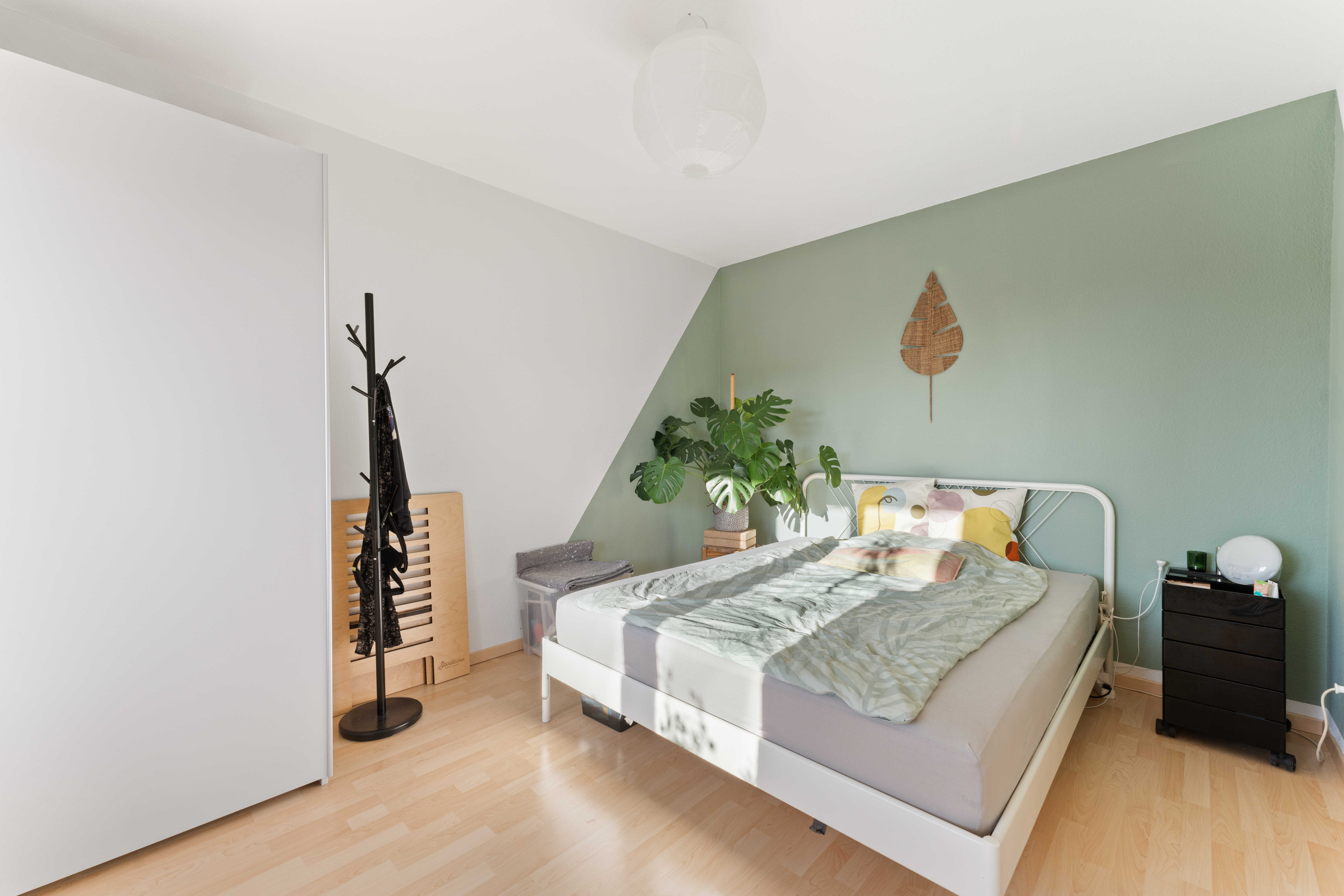 Helles Schlafzimmer mit grün gestrichener Wand, Doppelbett und Zimmerpflanze neben dem Bett.