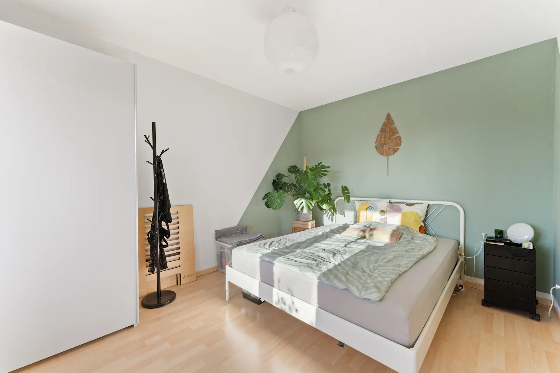 Helles Schlafzimmer mit grün gestrichener Wand, Doppelbett und Zimmerpflanze neben dem Bett.