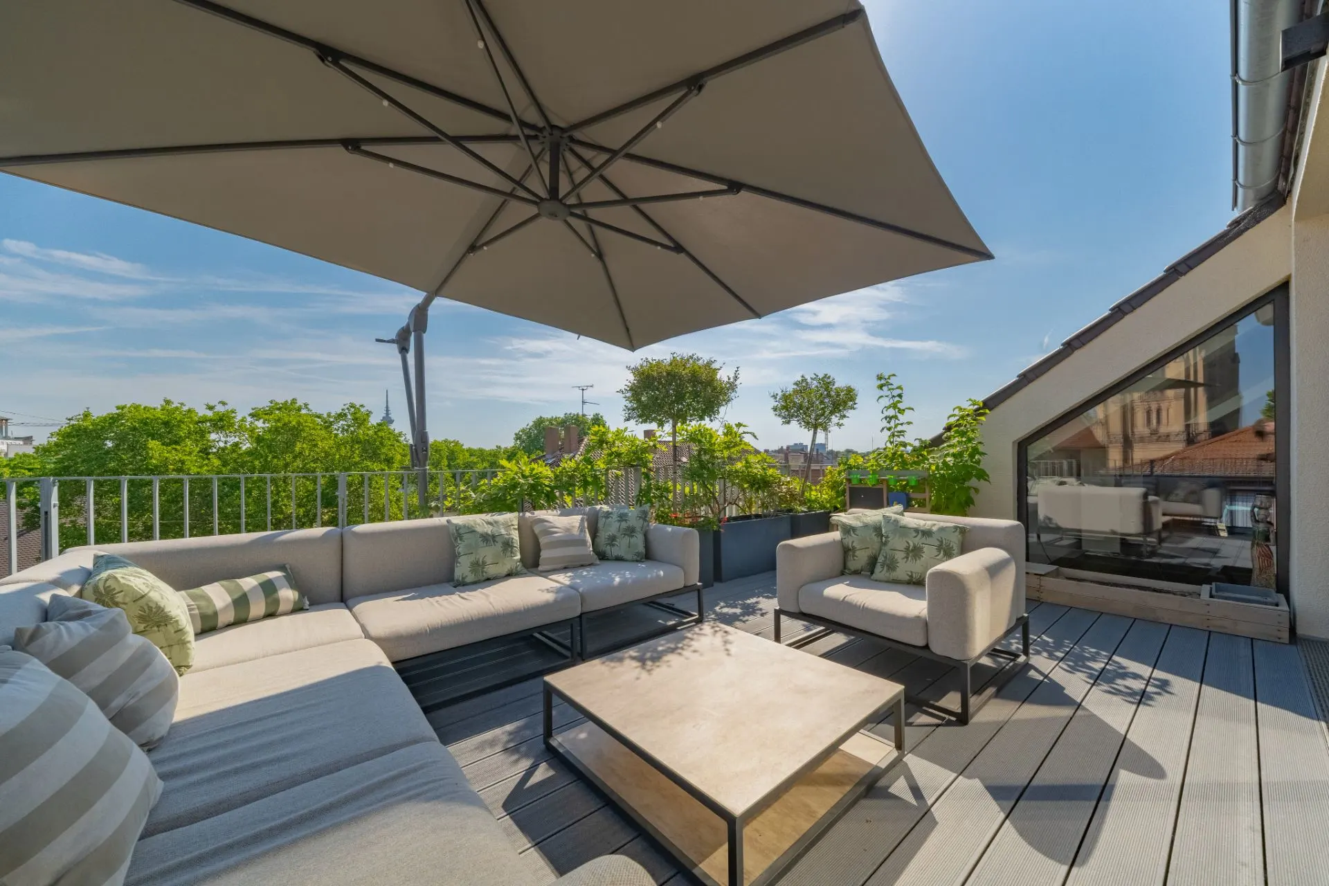 Große Terrasse mit Loungemöbeln, Kissen, Tisch und Sonnenschirm, umgeben von Pflanzen.