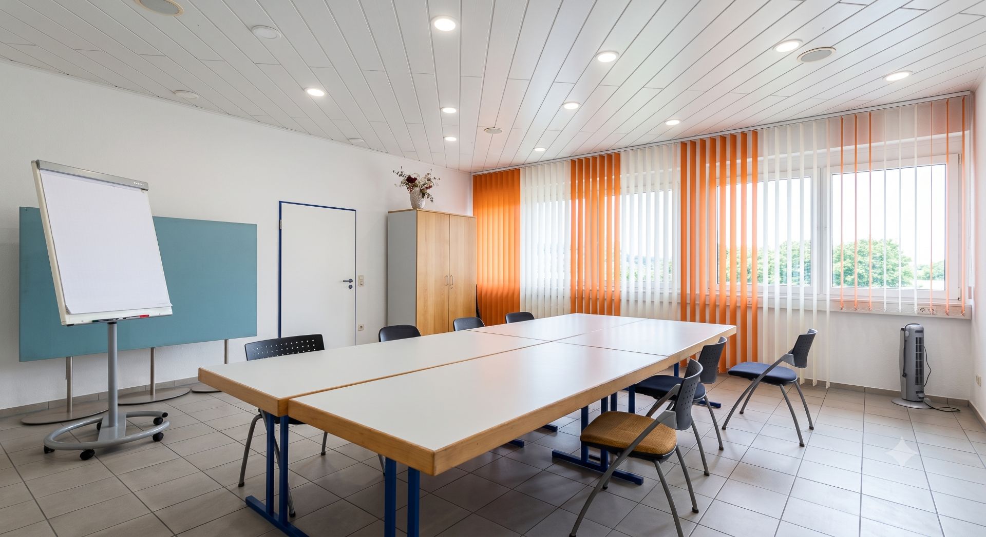 Heller Raum mit rechteckigem Tisch, mehreren Stühlen, Flipchart und großen Fenstern mit orange-weißen Vorhängen.