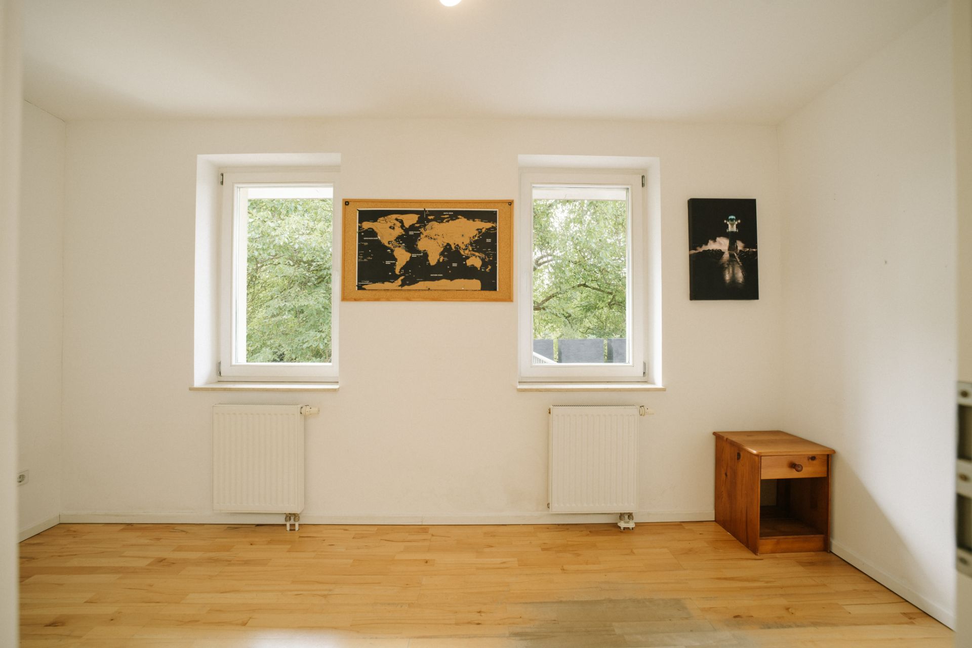 Helles Zimmer mit zwei Fenstern, zwei Heizkörpern, Holz-Nachttisch und zwei Bildern an der Wand.