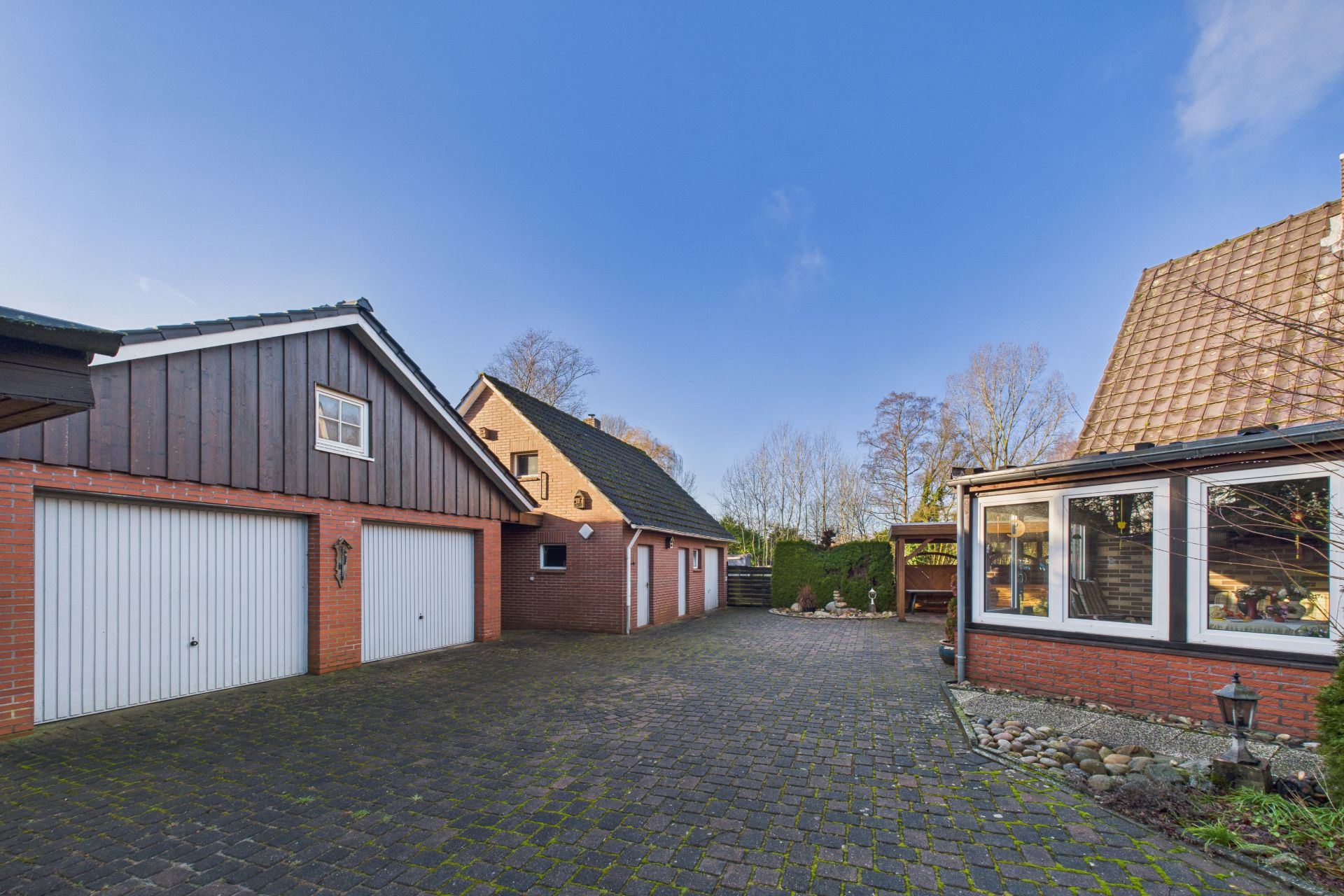 Gepflasterter Hof mit Garage, Wohnhaus und Wintergarten bei klarem Himmel.