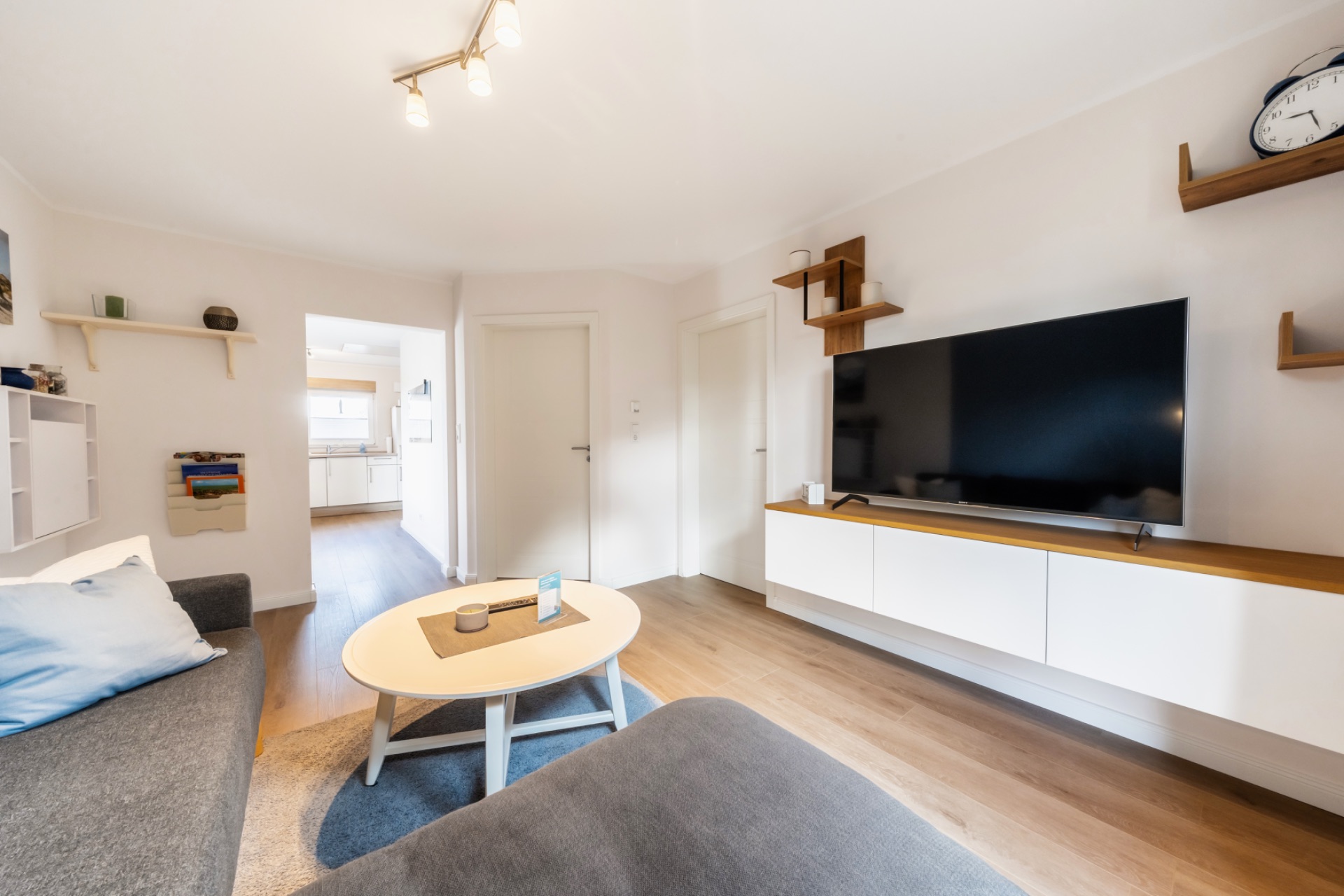 Helles Wohnzimmer mit grauem Sofa, rundem Tisch und großem Fernseher an der Wand.