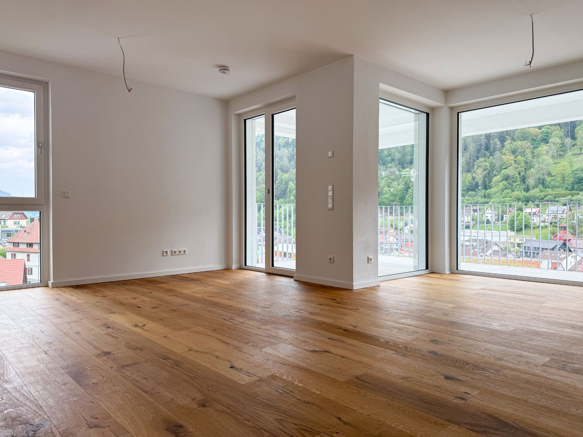 Leerer Wohnraum mit Holzfußboden, großen Fenstern und Blick auf grüne Hügel.