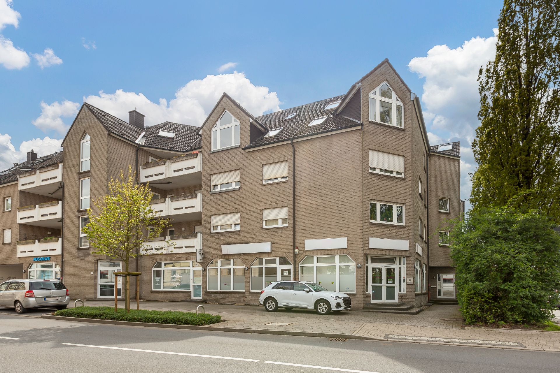 Mehrstöckiges Wohnhaus mit Balkonen, geparkten Autos und Bäumen an einer Straße.