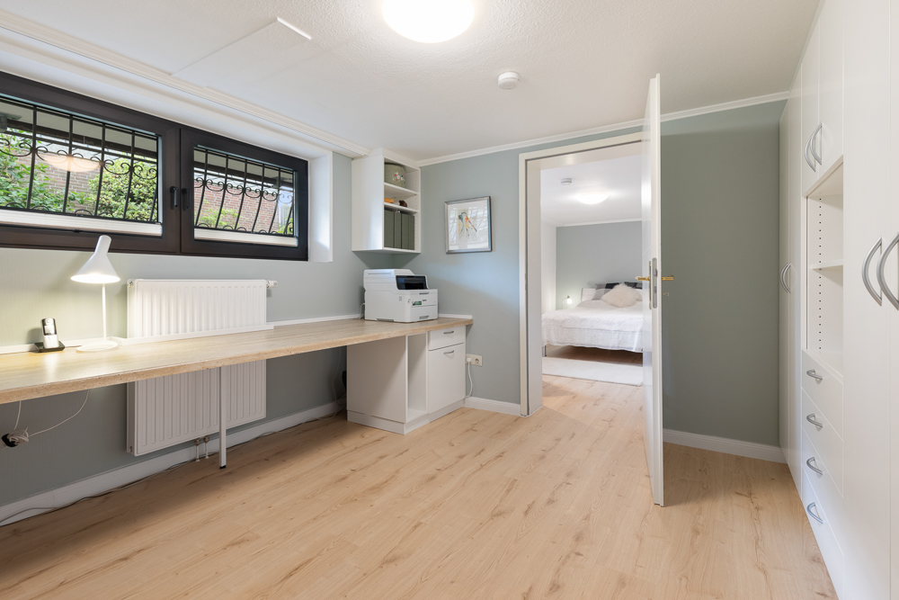 Heller Arbeitsraum mit langem Schreibtisch, Regal, Drucker und Blick in ein Schlafzimmer.
