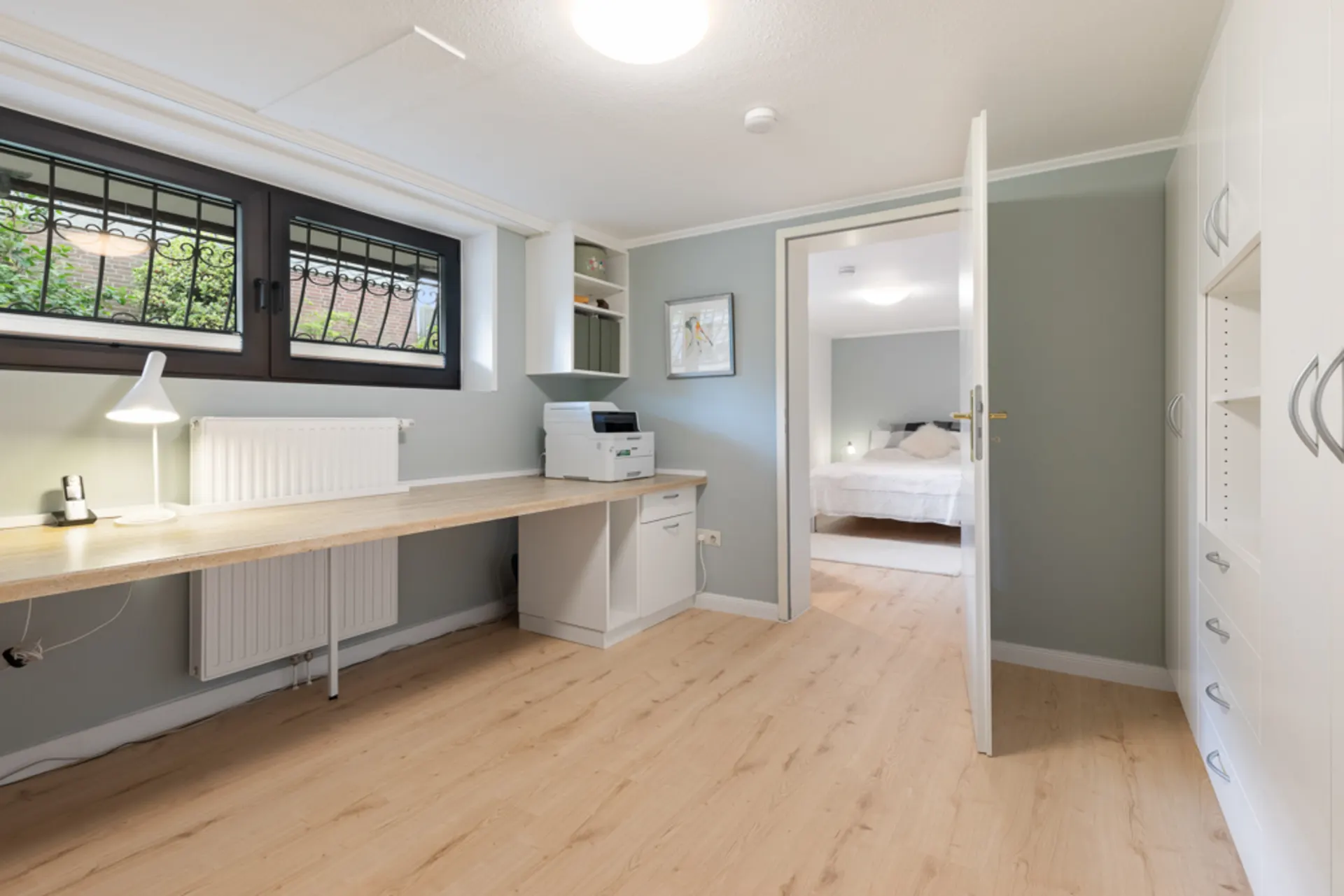 Heller Arbeitsraum mit langem Schreibtisch, Regal, Drucker und Blick in ein Schlafzimmer.