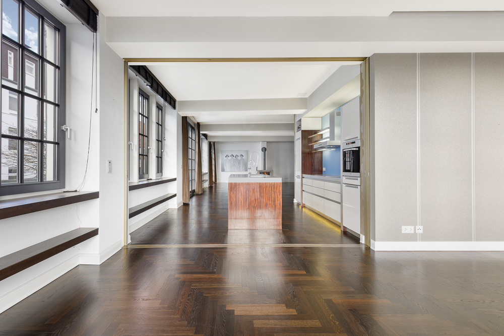 Großer, heller Raum mit Holzfußboden und moderner offener Küche, Fenster an der linken Seite.