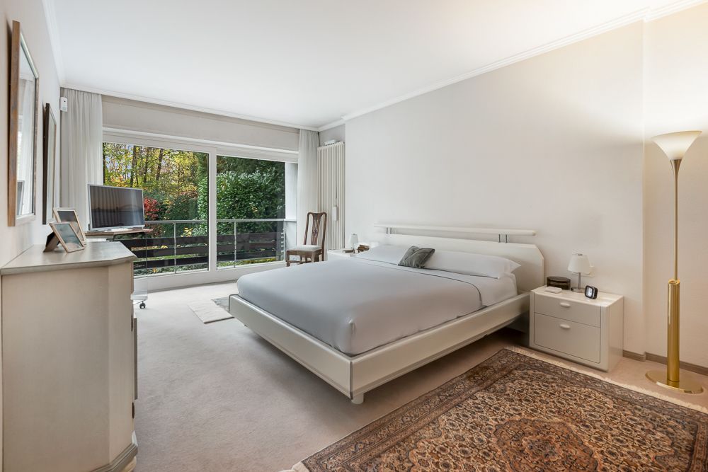 Helles Schlafzimmer mit Doppelbett, Teppich, Kommode, Fernseher und großem Fenster mit Blick ins Grüne.