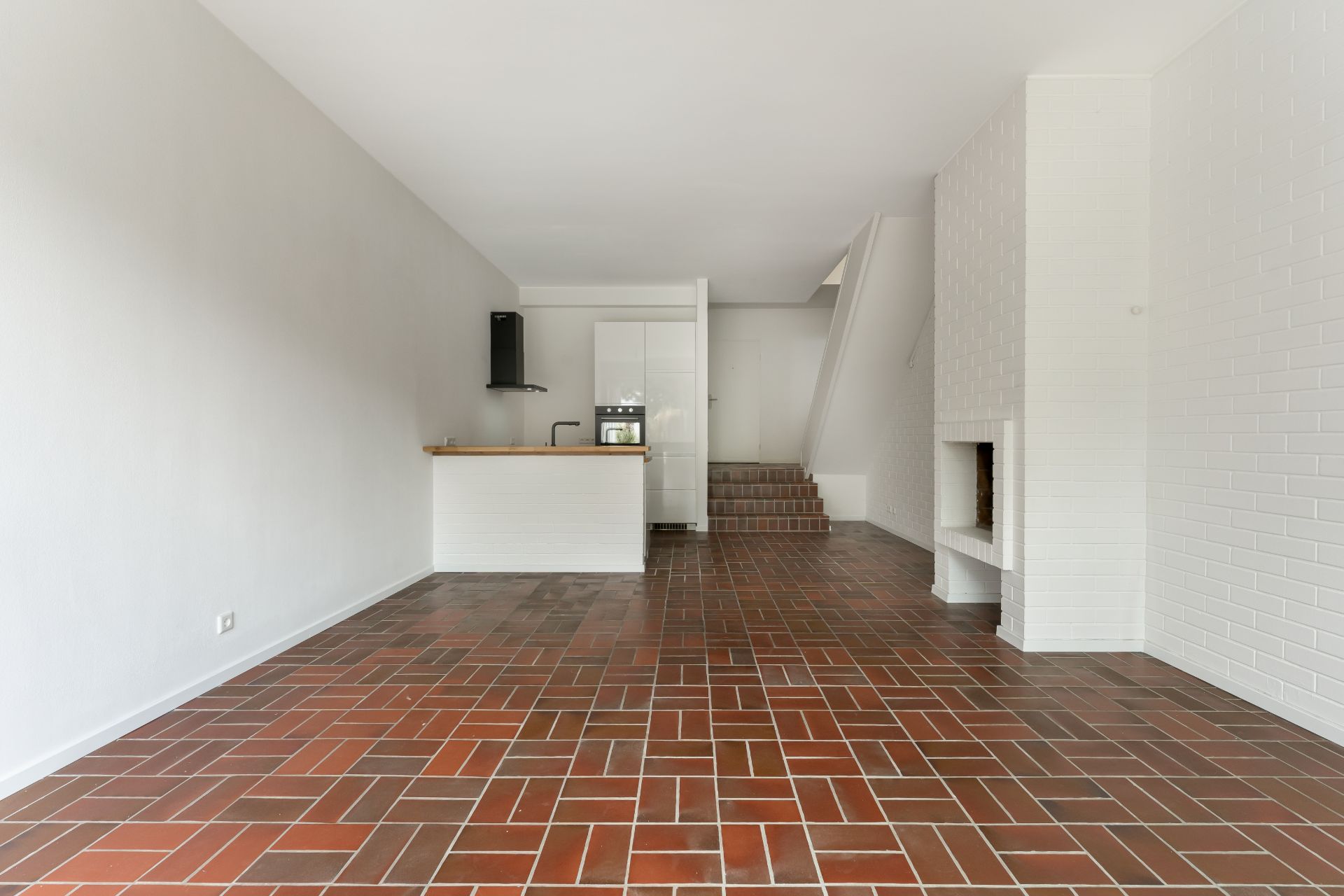 Leerer Wohnraum mit gemustertem Fliesenboden, offener Küche, Kamin und Treppe.